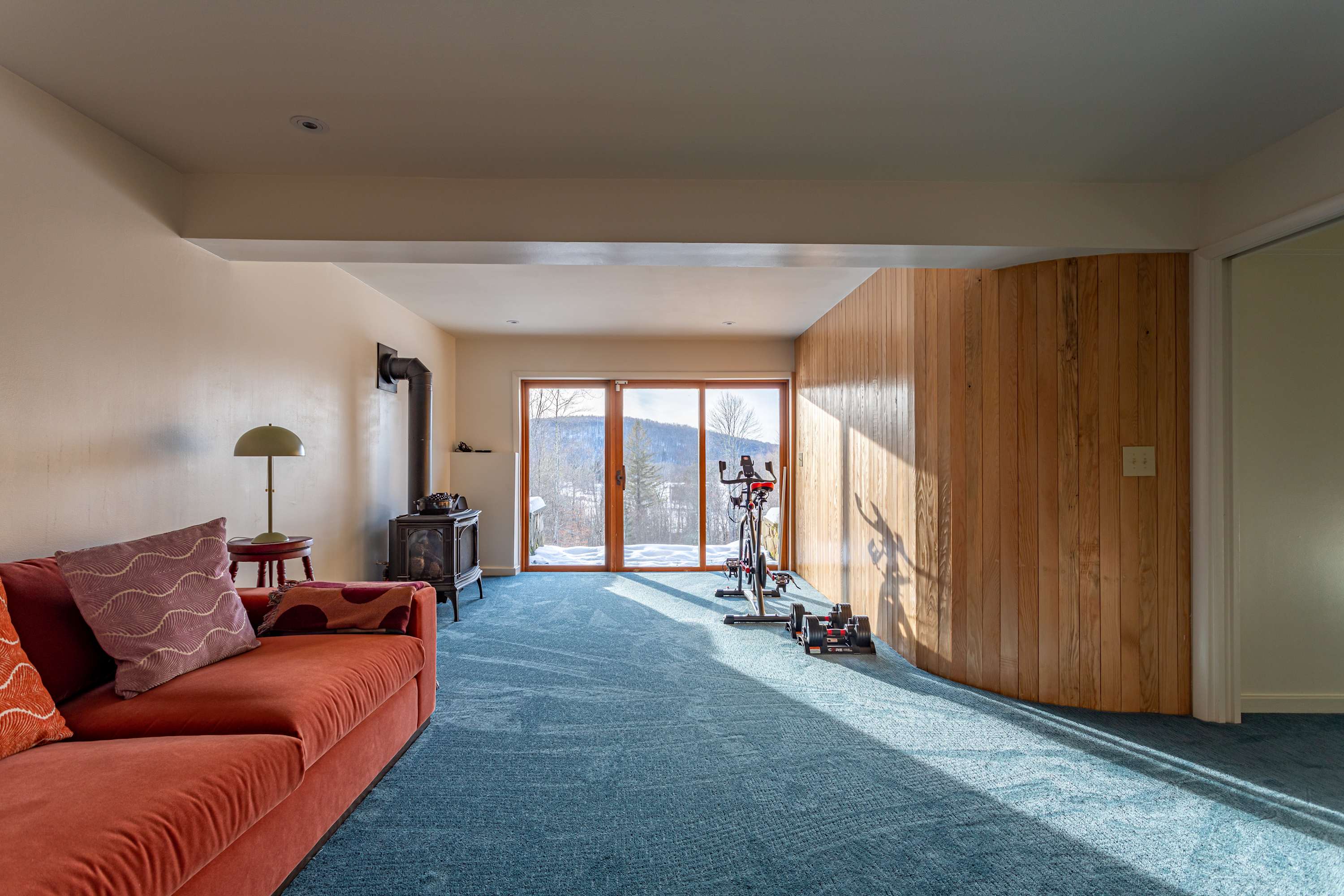 A spacious living area combines relaxation and fitness, featuring a red sofa, a black wood-burning stove, and an exercise bike. Large glass sliding doors offer panoramic views of a snowy mountain landscape, complemented by a wood-paneled wall.