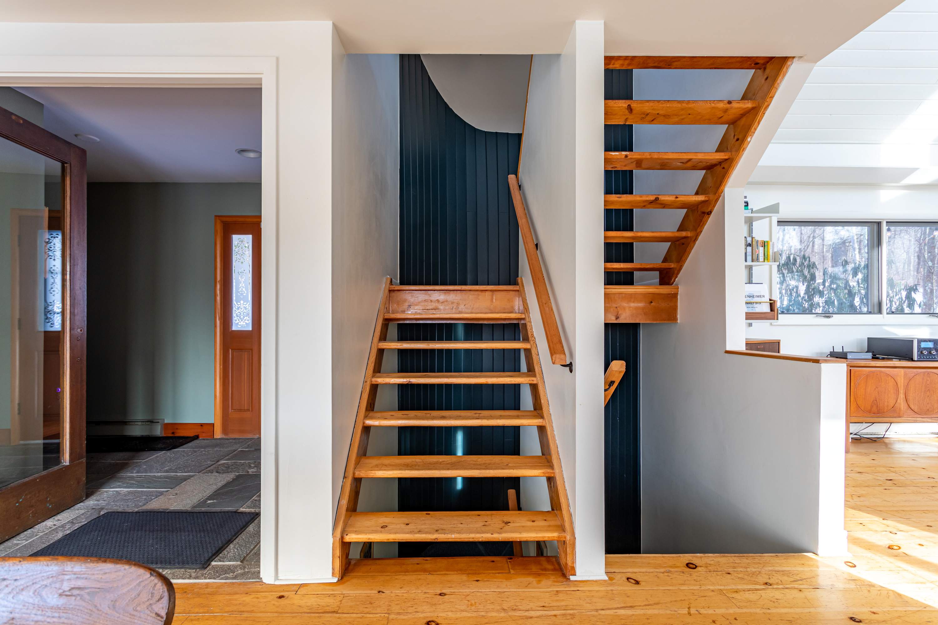 A welcoming foyer area featuring a wooden staircase with open risers leading to upper and lower levels. The entrance has stone tile flooring, transitioning to light wood floors on the upper landing.