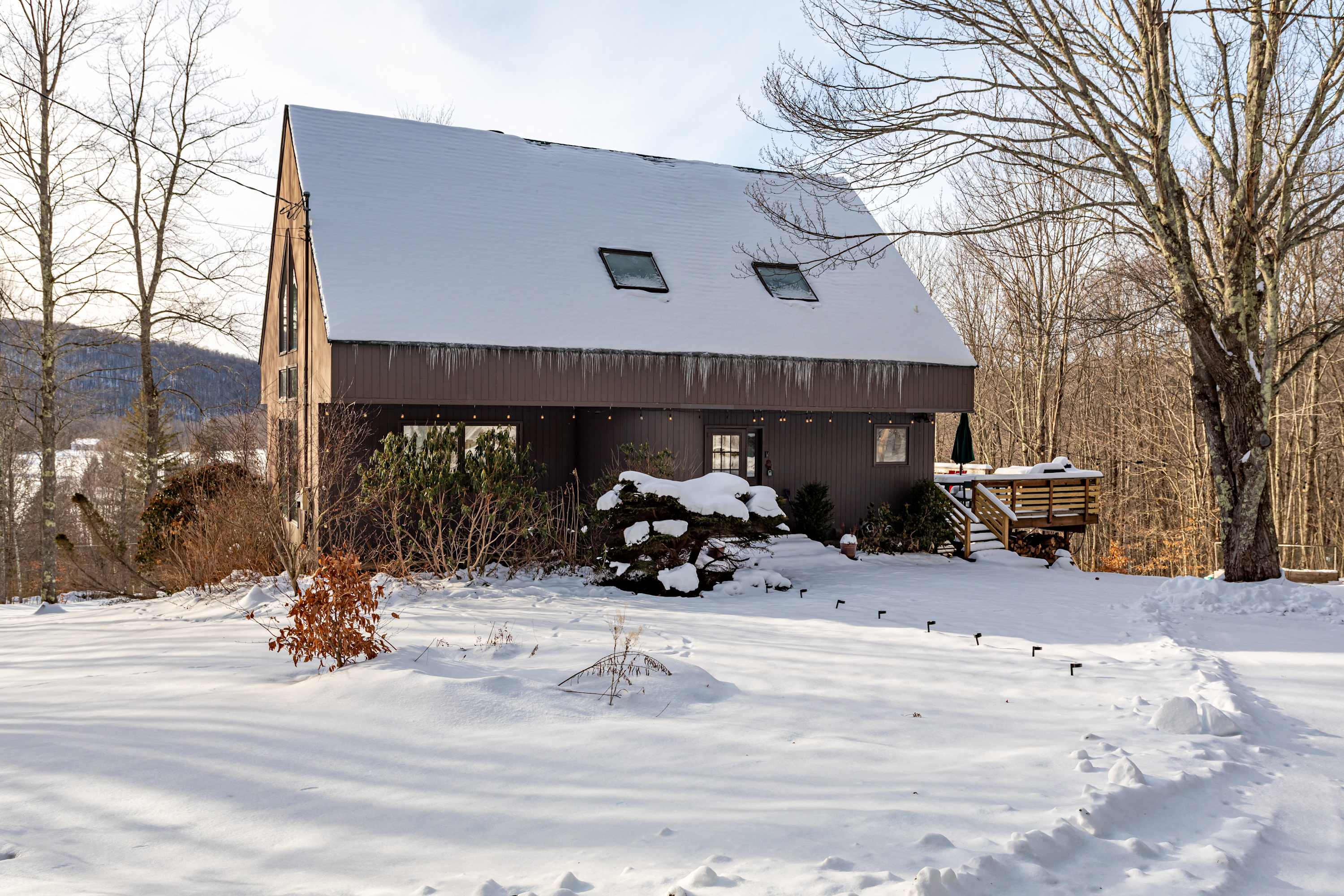 The side exterior of a brown A-frame house with a snow-covered roof and ground. Skylights are visible on the roof, and a wooden deck extends from the house, surrounded by bare trees under a bright sky.