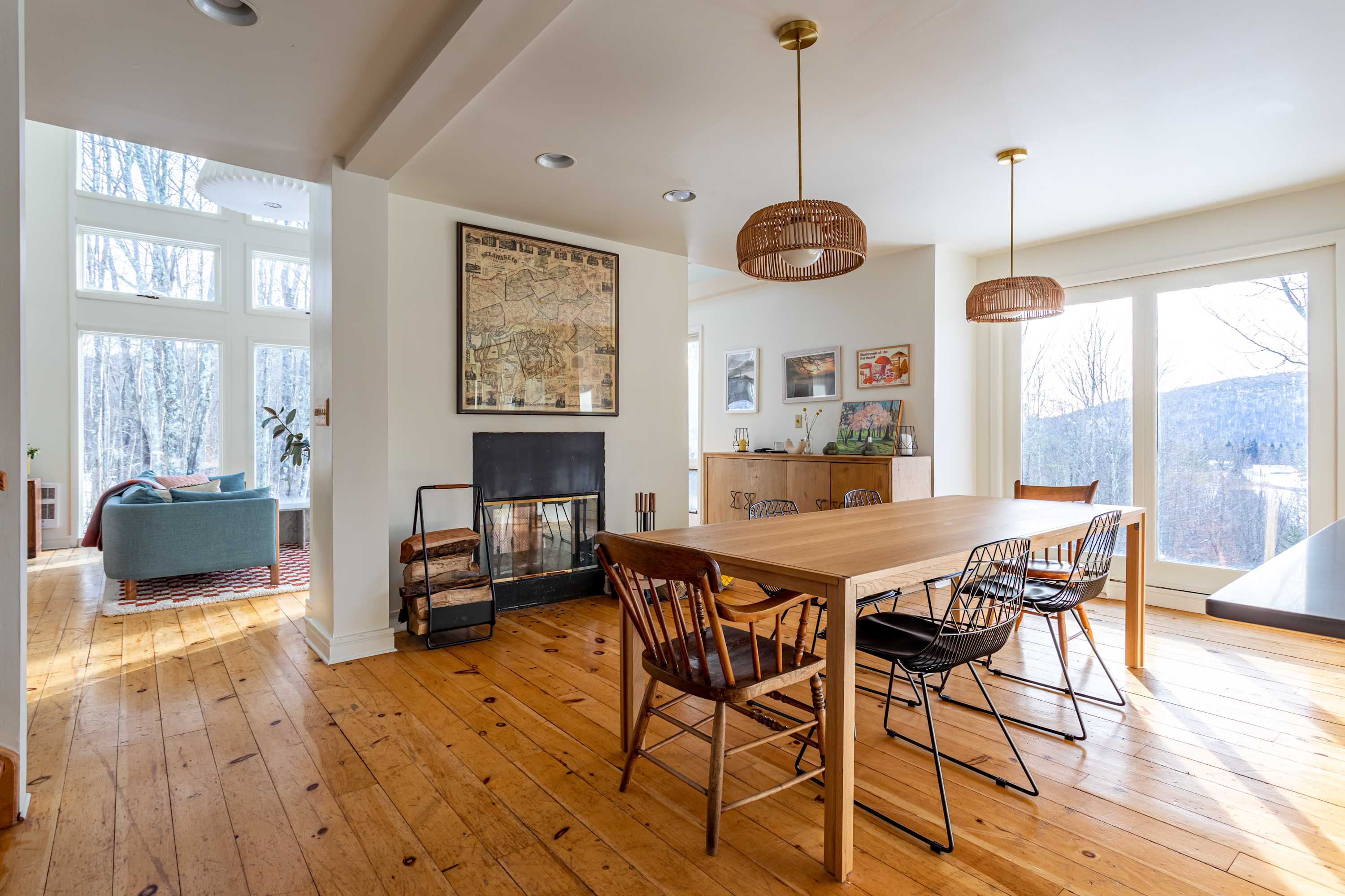 A bright dining area with light hardwood floors features a long wooden dining table surrounded by a mix of chairs, a modern fireplace, and large windows offering views of the winter forest.