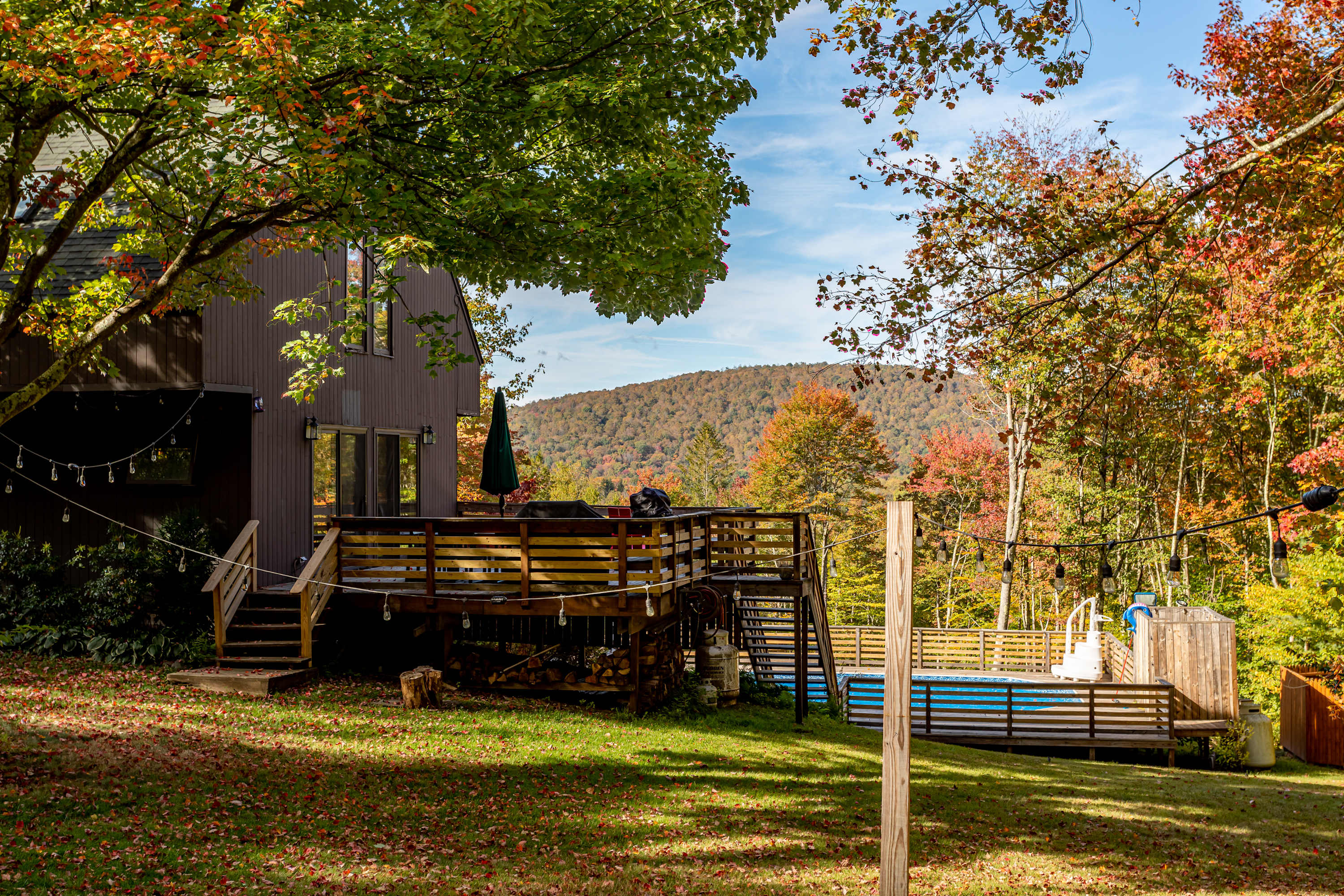 The back of the brown house features a multi-level wooden deck with outdoor seating and a grill, overlooking an above-ground pool and a vibrant autumn landscape with rolling hills.