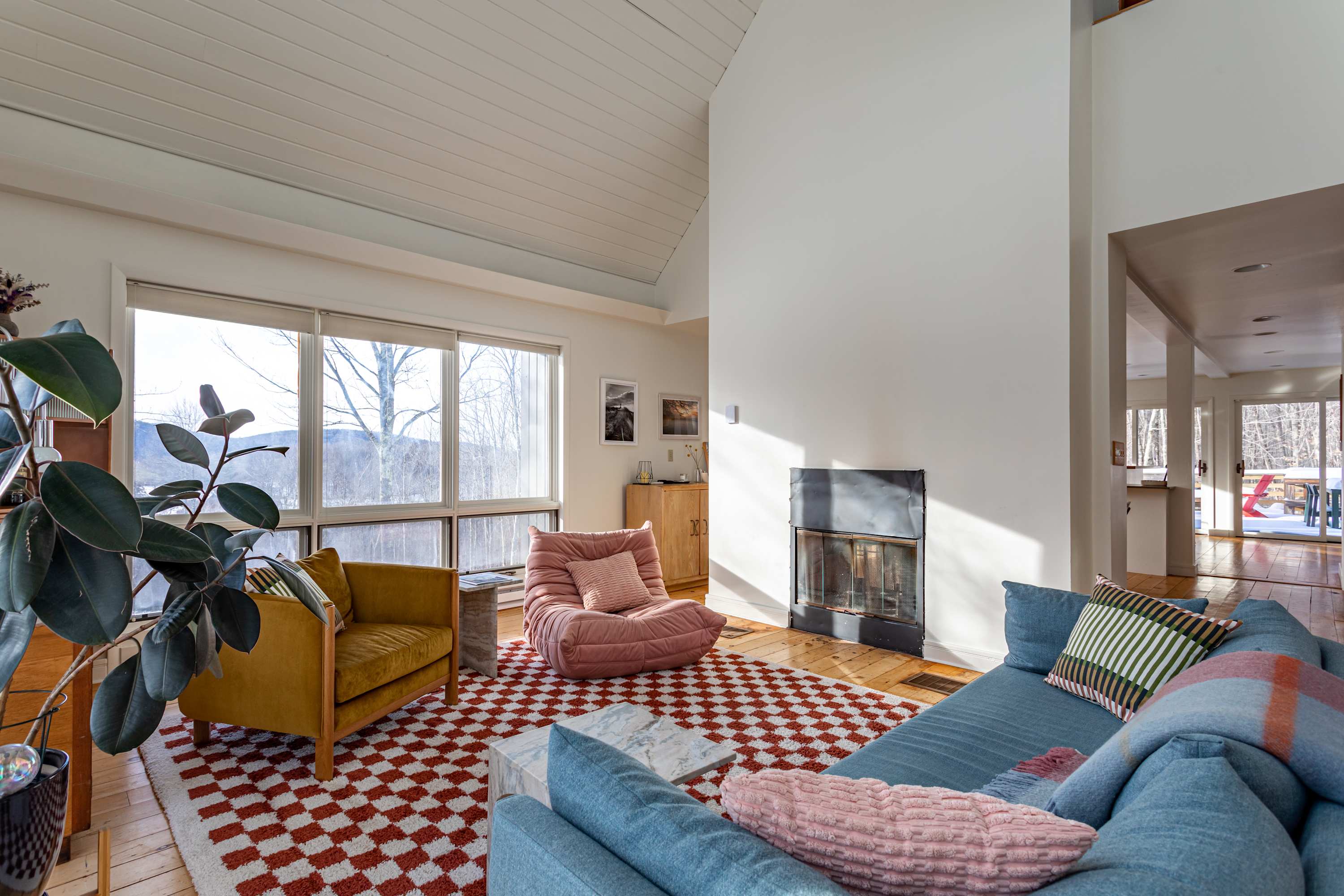 A cozy living room with a vaulted white ceiling and light hardwood floors features a modern fireplace, a blue sofa, a pink lounge chair, and a yellow armchair, all arranged on a red and white checkered rug. Large windows offer views of a winter landscape.