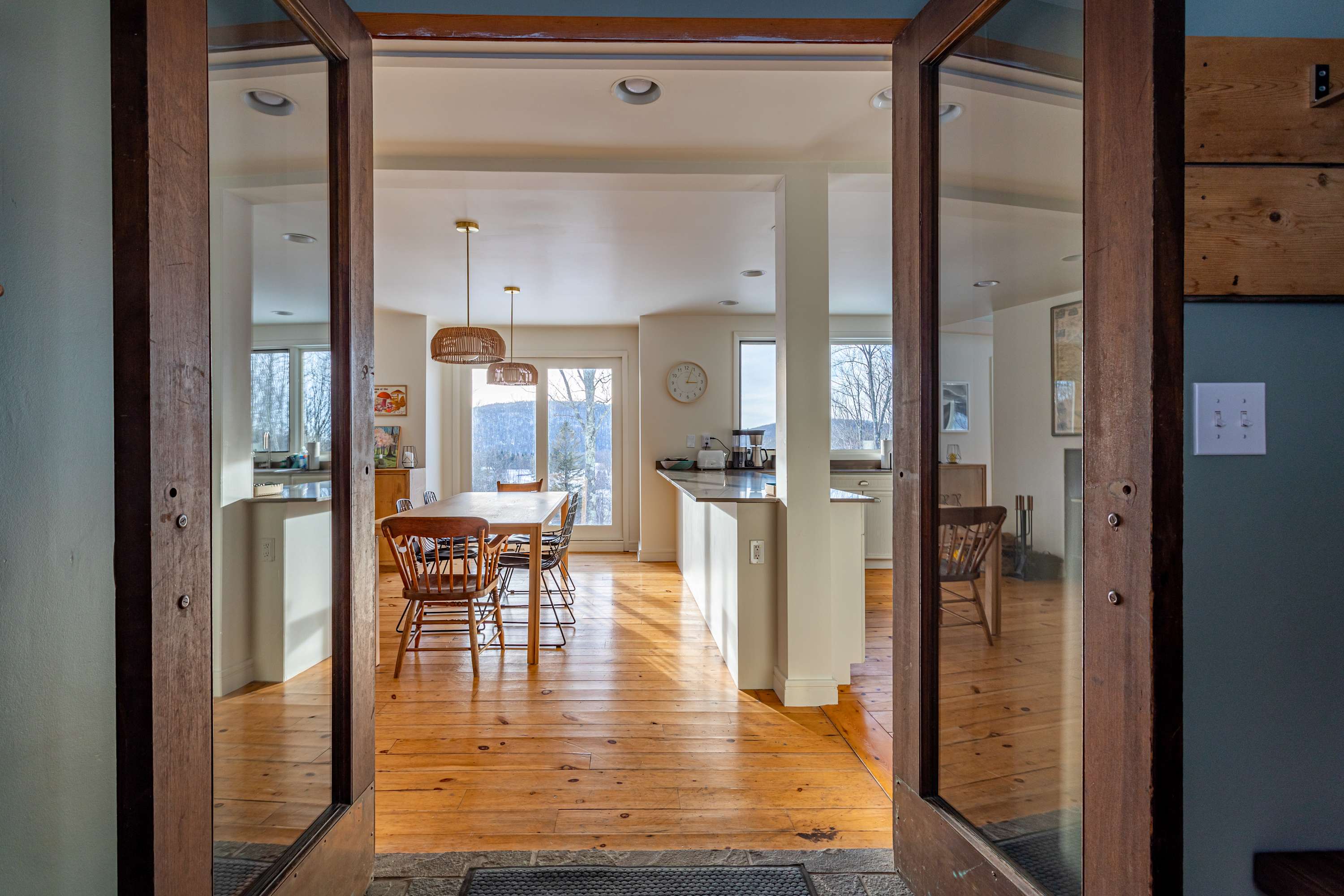 A welcoming entryway with double glass-paned doors opening into an open-concept dining and kitchen area. The space features warm hardwood floors and bright natural light from large windows.