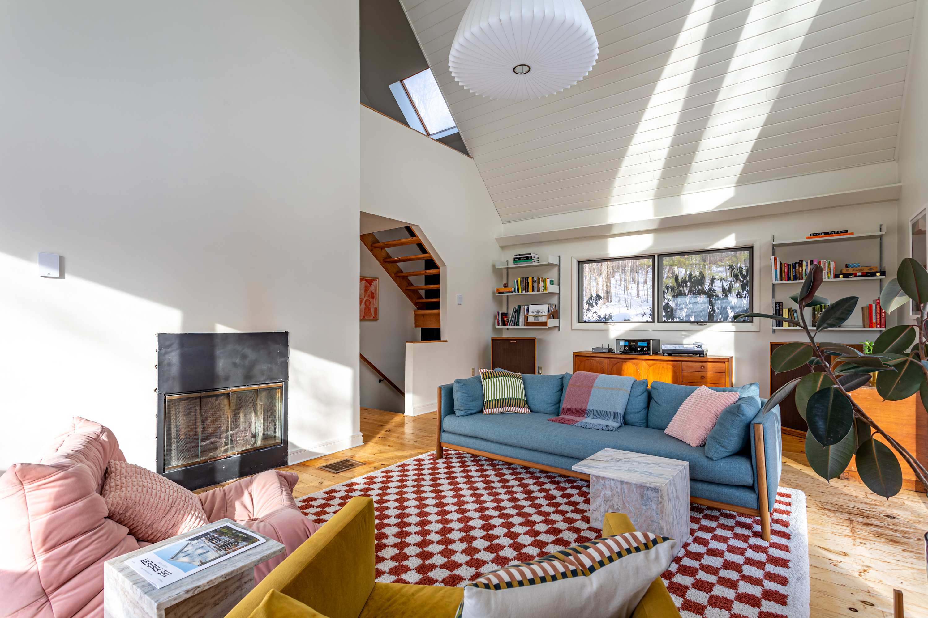 A spacious living room with a black fireplace and a light blue sofa, set on a red and white checkered rug. A staircase leads to an upper level, and large windows offer views of the snowy outdoors.