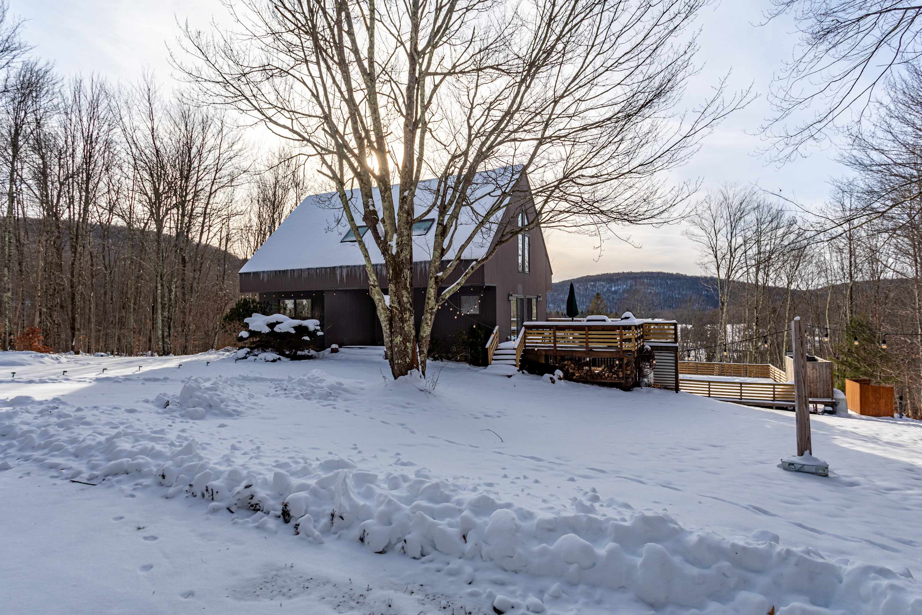 The front exterior of a brown A-frame house is seen through a sparse screen of bare trees, with a wide expanse of snow-covered yard in the foreground. A wooden deck is visible to the right, and distant hills complete the winter scene.