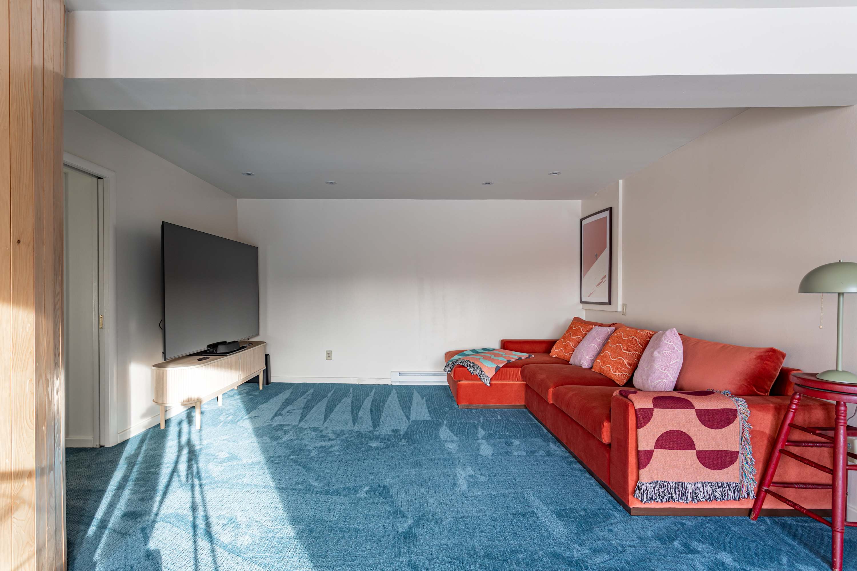 A bright living room features a large red sectional sofa with colorful throw pillows and blankets, facing a wall-mounted TV on a light wood console. A vibrant blue patterned carpet covers the floor, with sunlight streaming in.