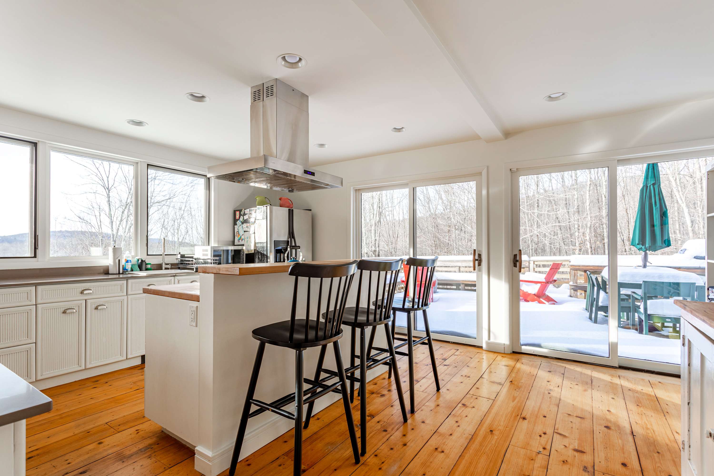 A modern kitchen featuring a central island with black bar stools and stainless steel appliances, including a range hood. Large sliding glass doors lead to an outdoor deck with snowy views.
