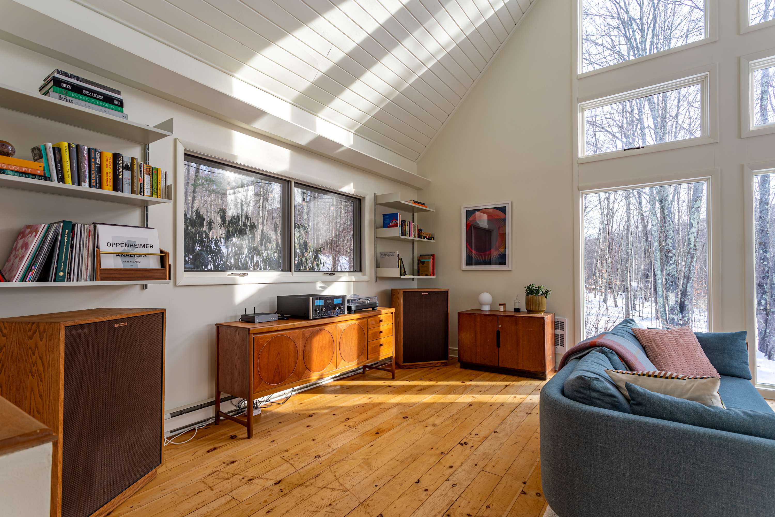 A bright living room corner featuring a vintage audio system with large speakers and a wooden credenza. Built-in shelves display books, and large windows provide views of the snowy forest.