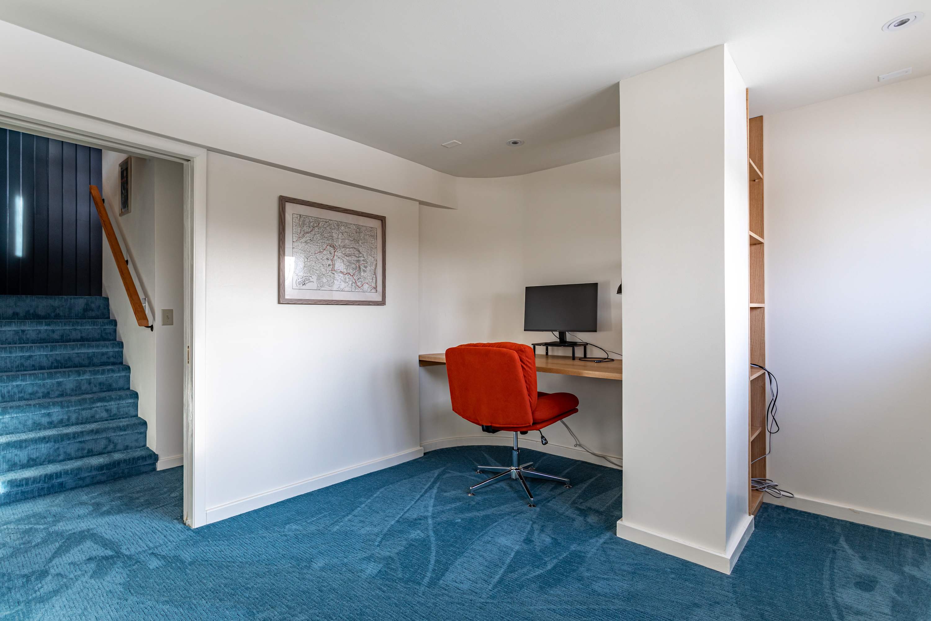A dedicated office space featuring a built-in wooden desk with a computer monitor and an orange ergonomic chair. The area has clean white walls, blue carpet, and is conveniently located near a carpeted staircase.
