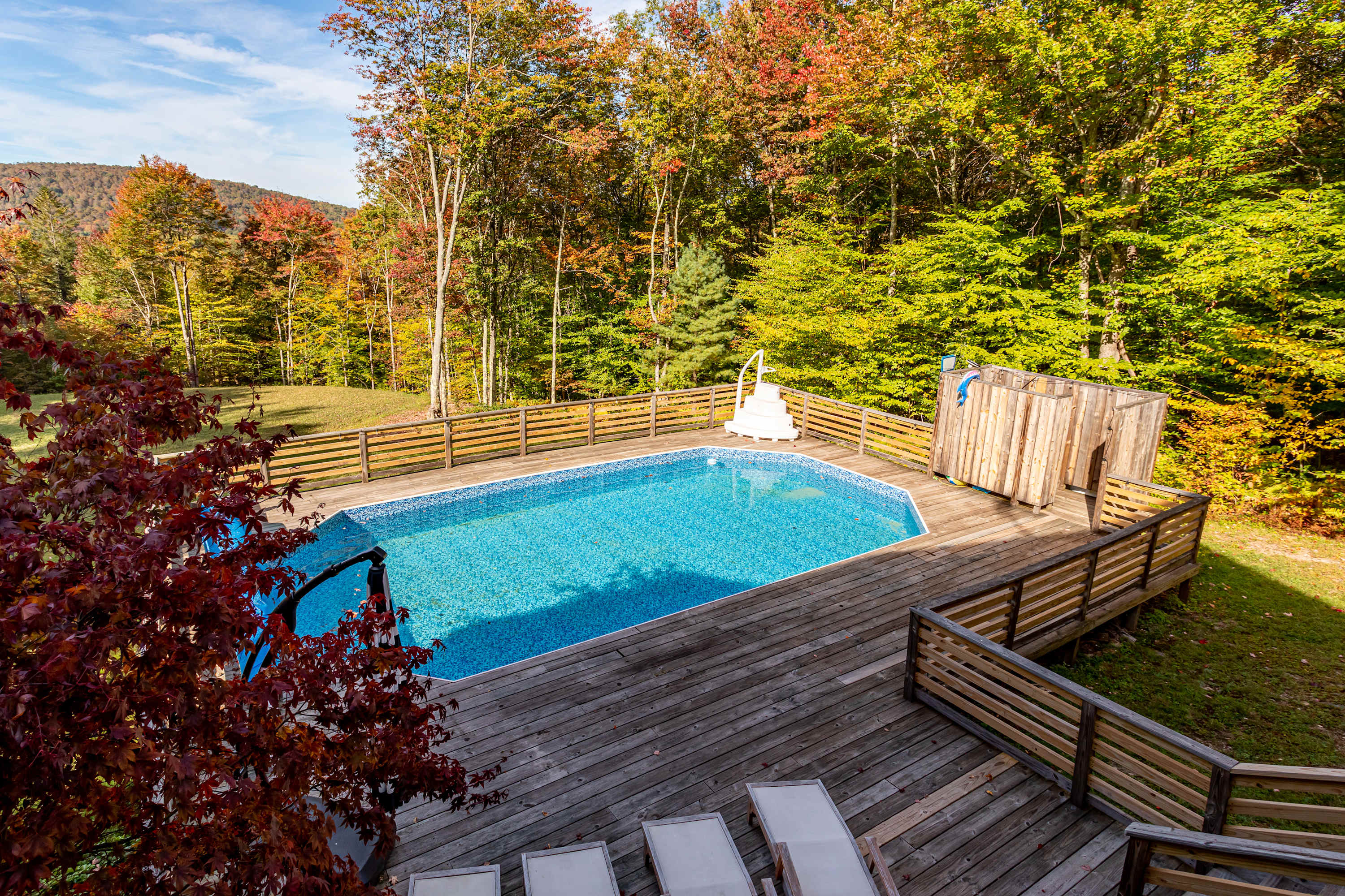 An aerial perspective showcases the blue swimming pool nestled within a large wooden deck, bordered by a rustic wooden fence and vibrant autumn foliage, with a small red-leafed tree and an outdoor shower enclosure.