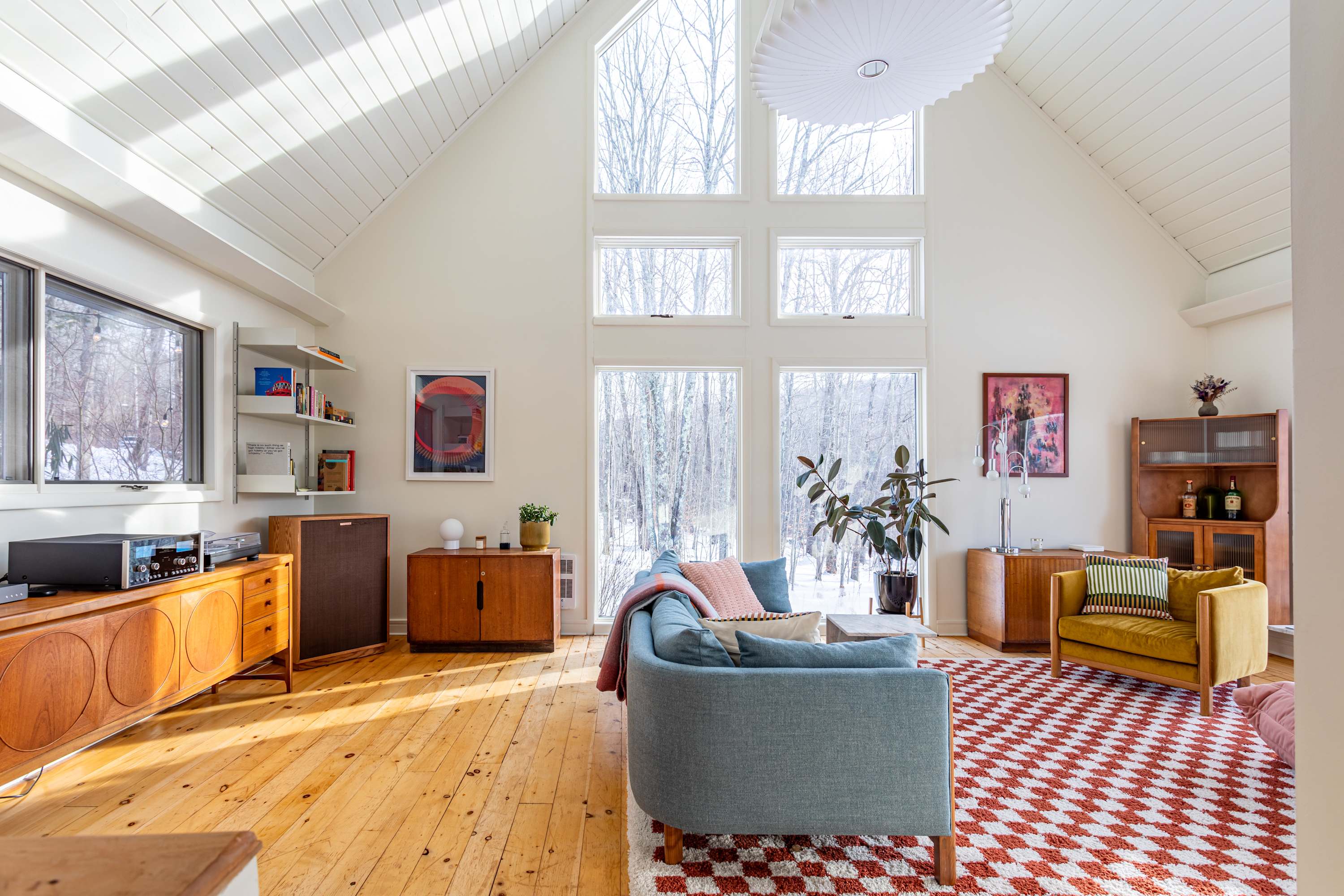 A wide view of the airy living room showcasing its vaulted ceiling, large windows, and a mix of mid-century modern furniture. A blue sofa and yellow armchair are arranged on a vibrant red and white checkered rug.