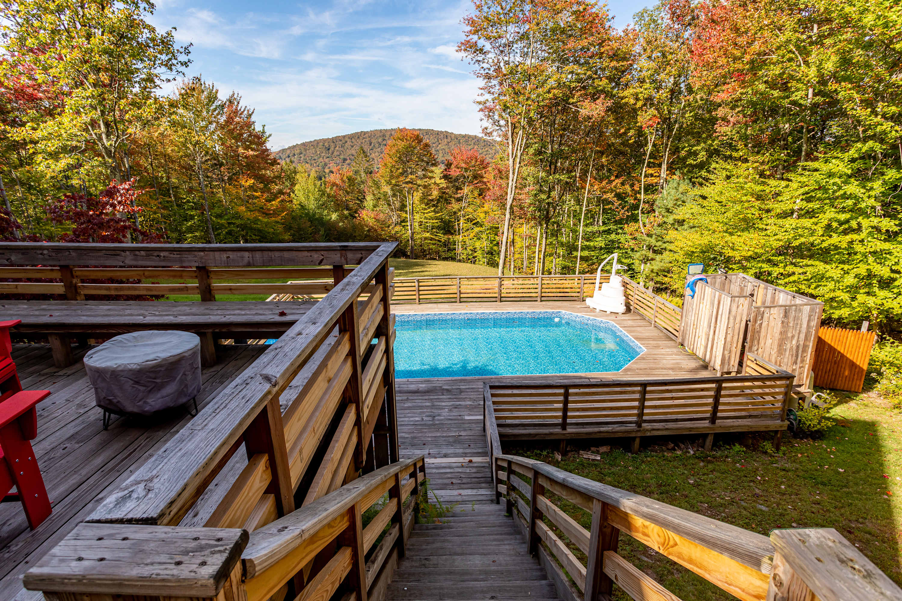 An elevated view captures a multi-level wooden deck with stairs leading down to a blue swimming pool, surrounded by a wooden fence and lush autumn trees, with distant mountains under a clear sky.