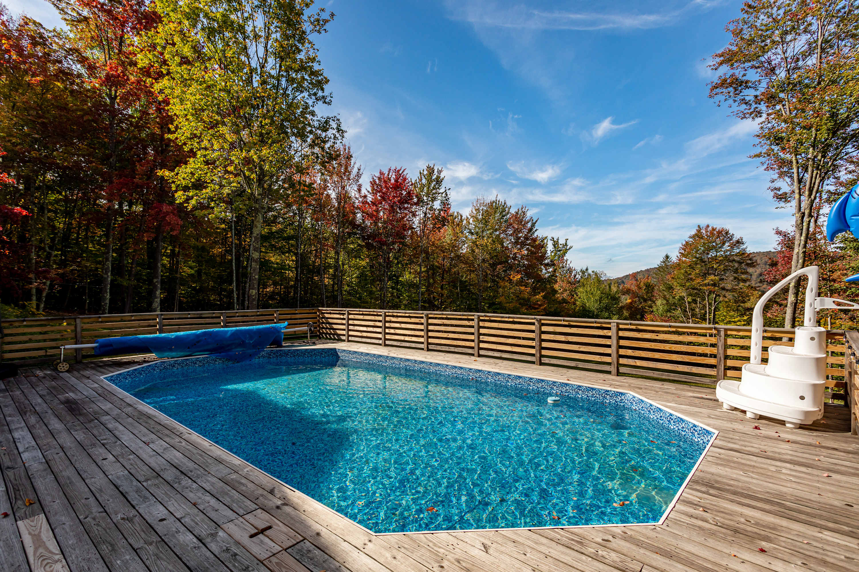 A clear blue swimming pool is surrounded by a wooden deck and a horizontal slatted fence, with vibrant autumn trees and a bright blue sky with white clouds in the background. A white pool ladder and a blue pool cover are visible.
