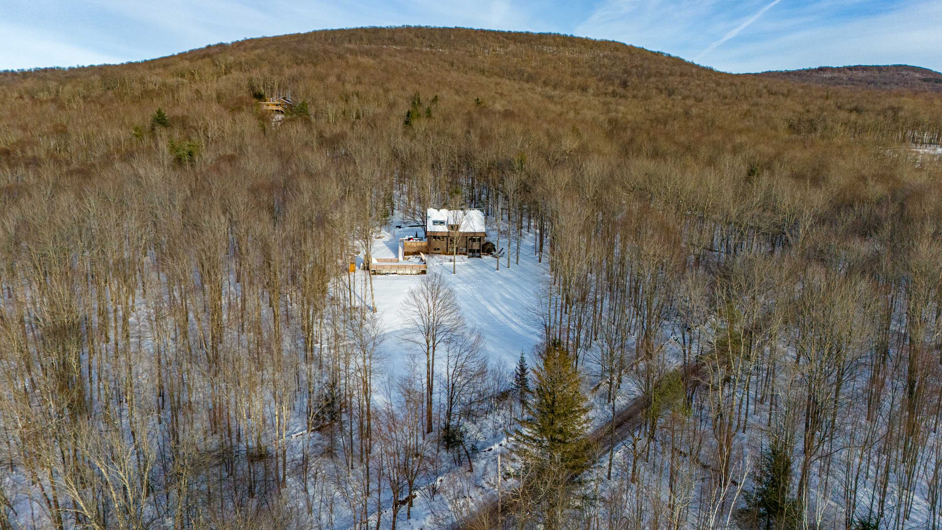 An aerial view captures the brown house and its surrounding snowy landscape, nestled within a dense forest of bare winter trees, highlighting its secluded mountain setting.