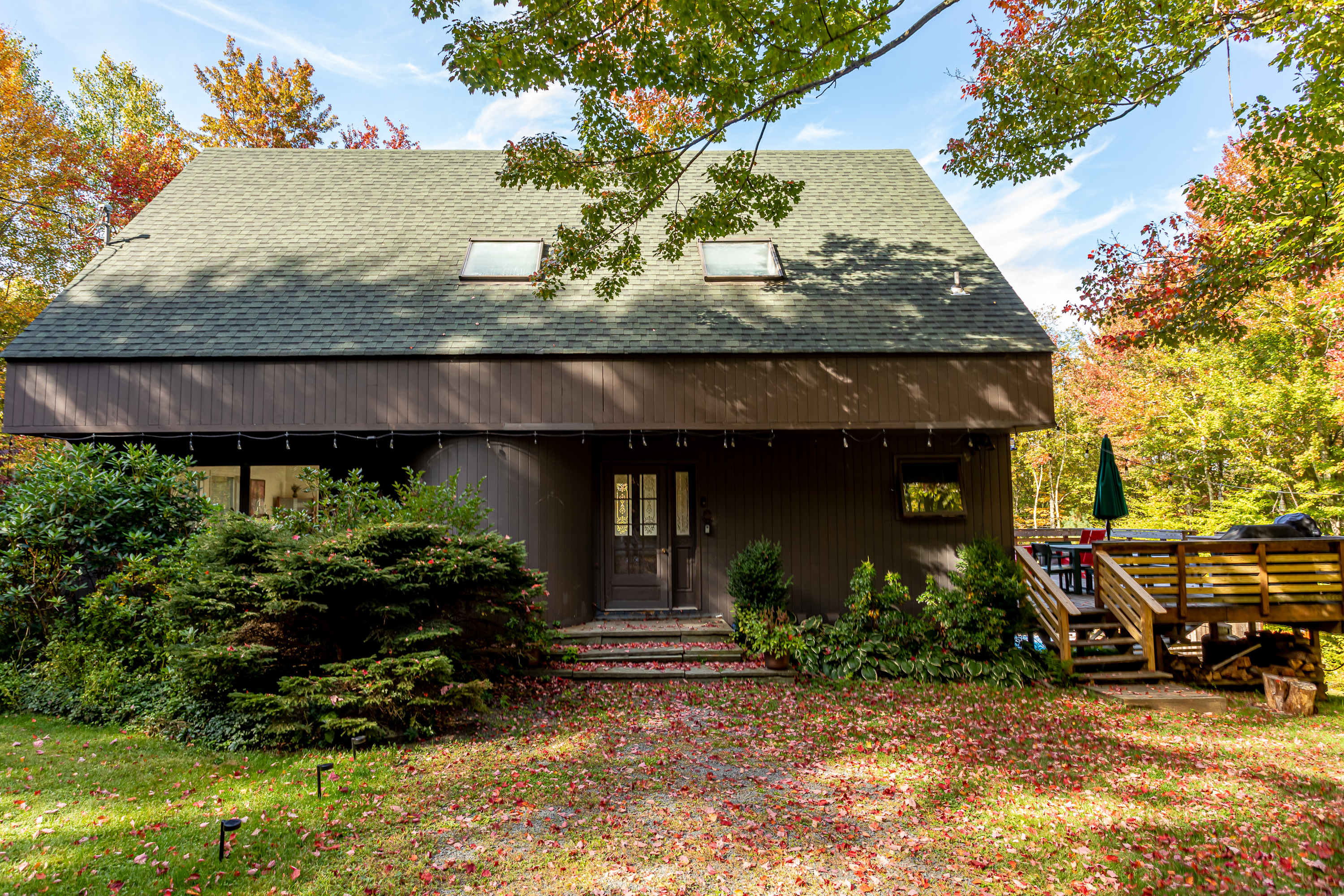The front facade of a brown, two-story house with a dark green roof and two skylights. The entrance features a dark wood door with glass panels, flanked by green bushes and red autumn leaves scattered on the ground.