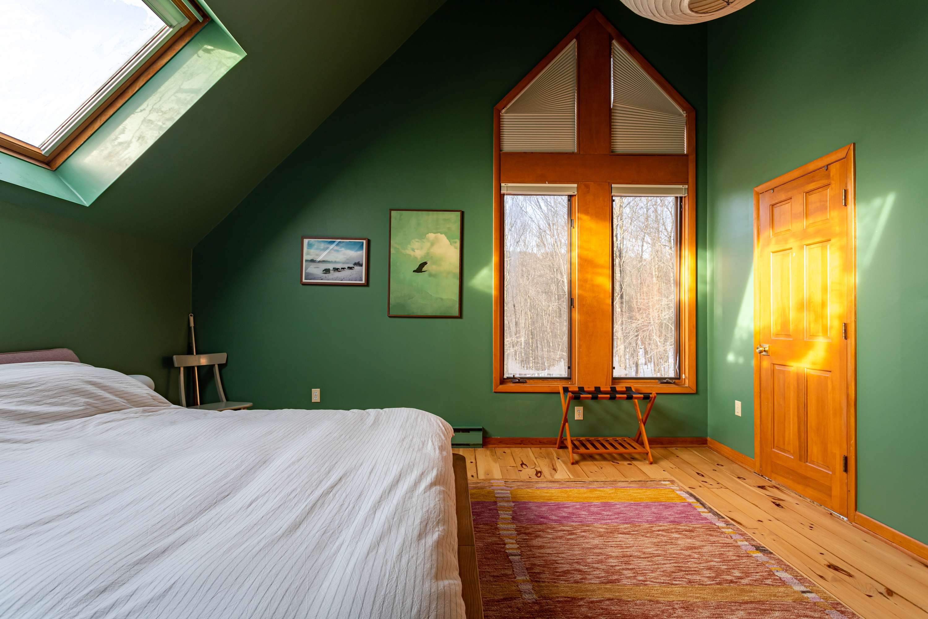 A cozy bedroom featuring striking dark green walls, light wood floors, and a large bed with crisp white bedding. A unique A-frame window and a skylight provide ample natural light and views of a winter forest.
