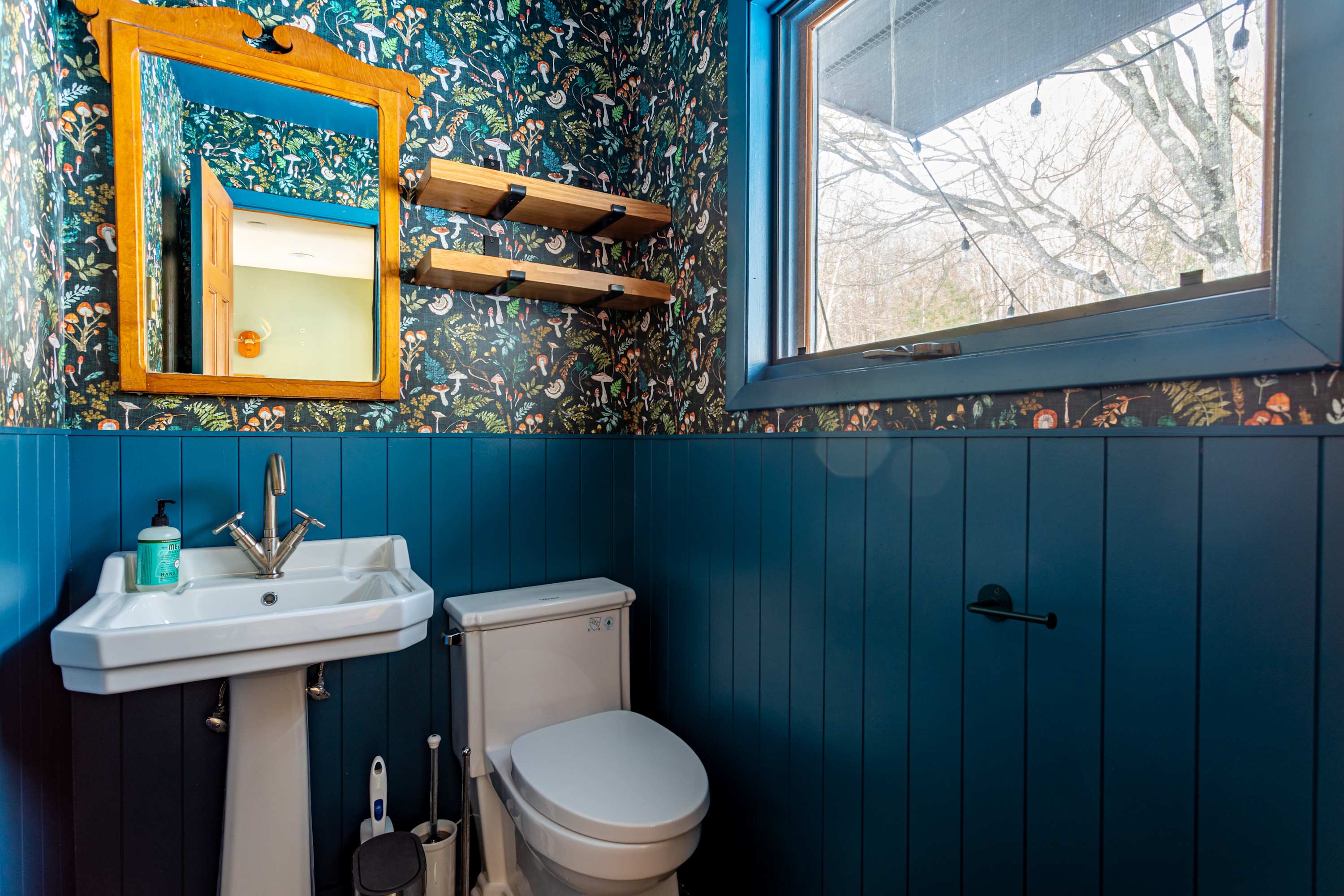A vibrant powder room features bold mushroom-patterned wallpaper above dark blue wainscoting. It includes a white pedestal sink with a vintage-style faucet, a toilet, and a window offering a glimpse of bare trees.