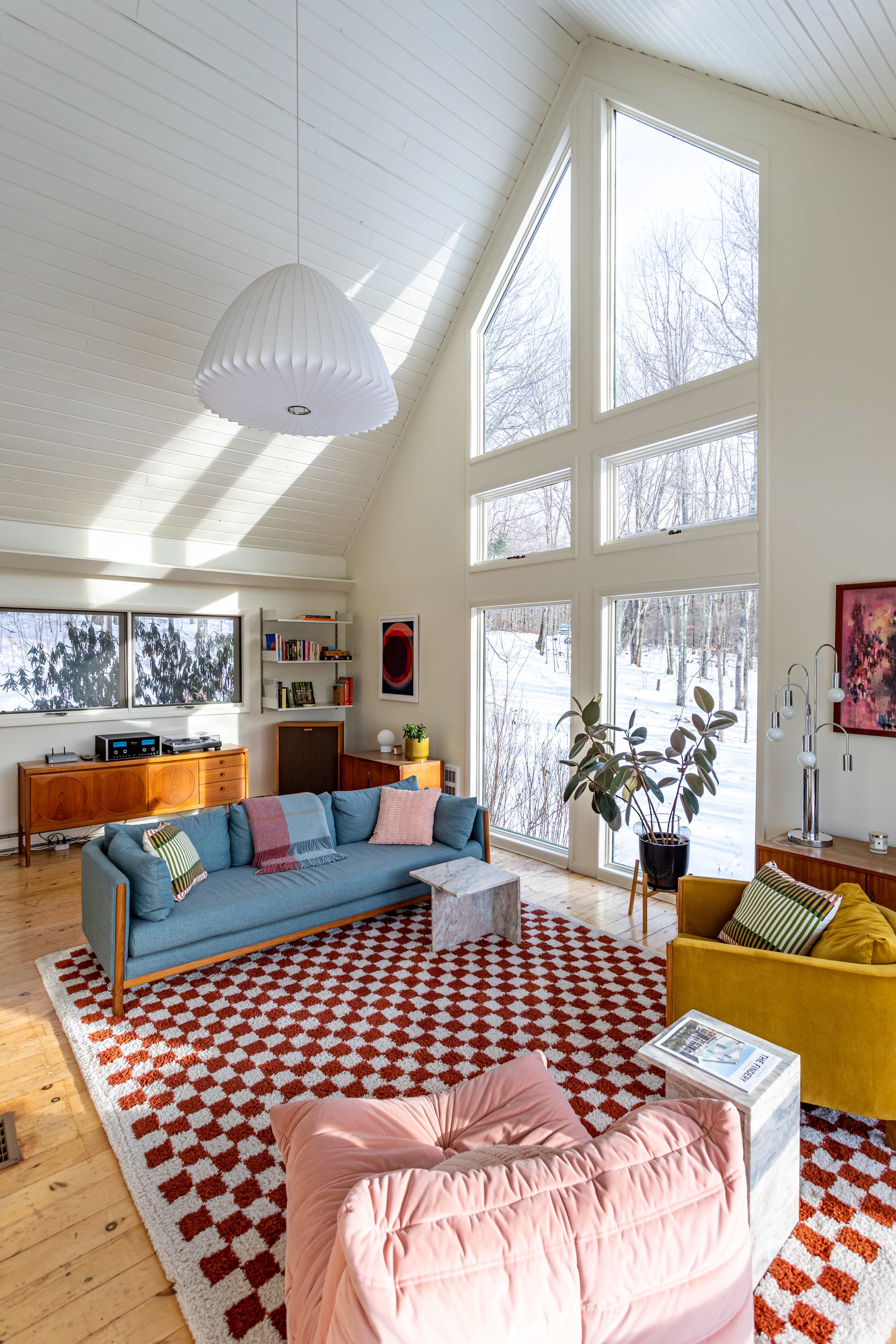 A bright, spacious living room with a high vaulted ceiling and large windows offering winter forest views. A blue sofa, yellow armchair, and pink pouf are arranged on a red and white checkered rug over light hardwood floors.
