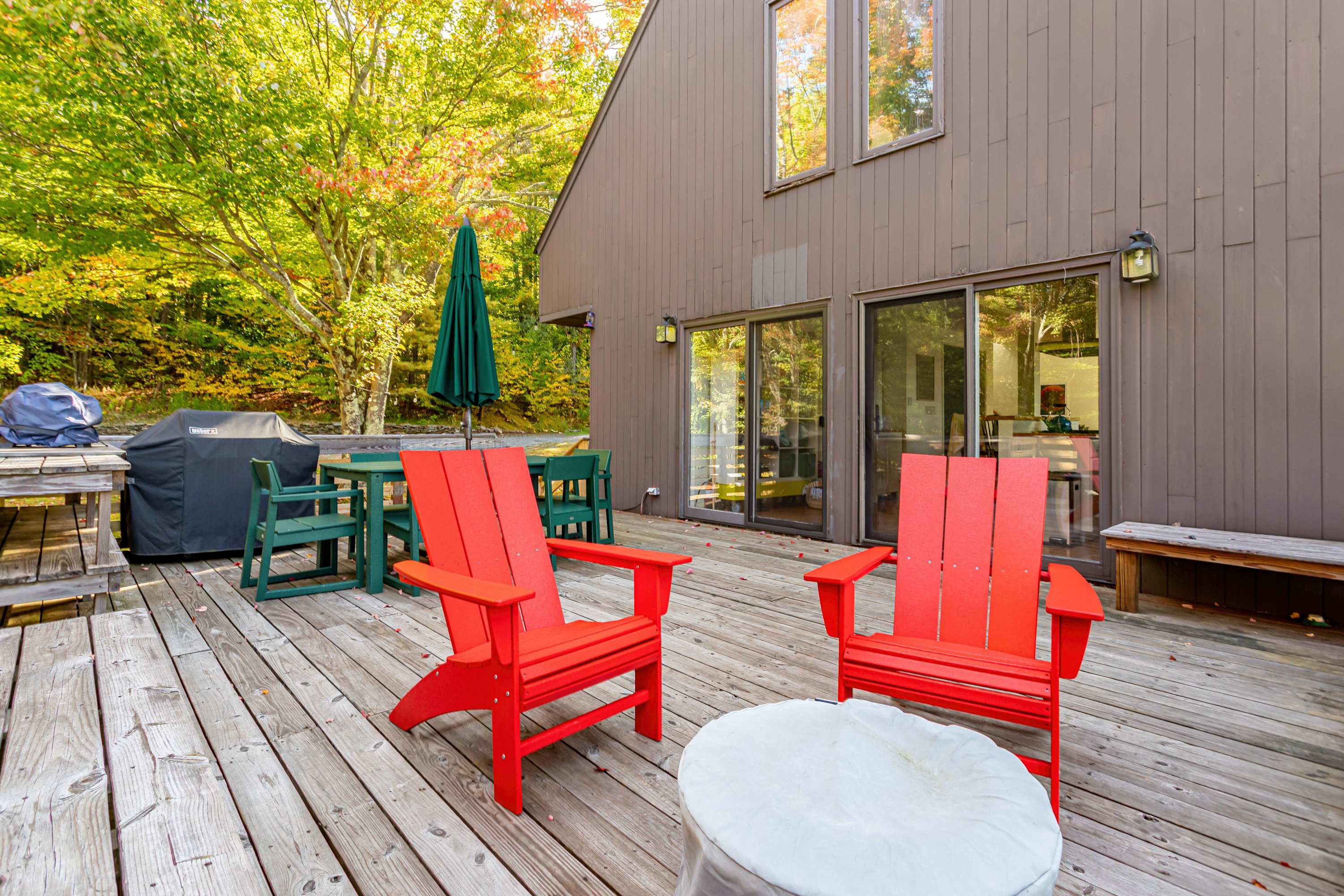 The expansive wooden deck offers a comfortable outdoor living space with red Adirondack chairs, a covered fire pit, and a dining set, adjacent to the house with large sliding glass doors and surrounded by lush trees.
