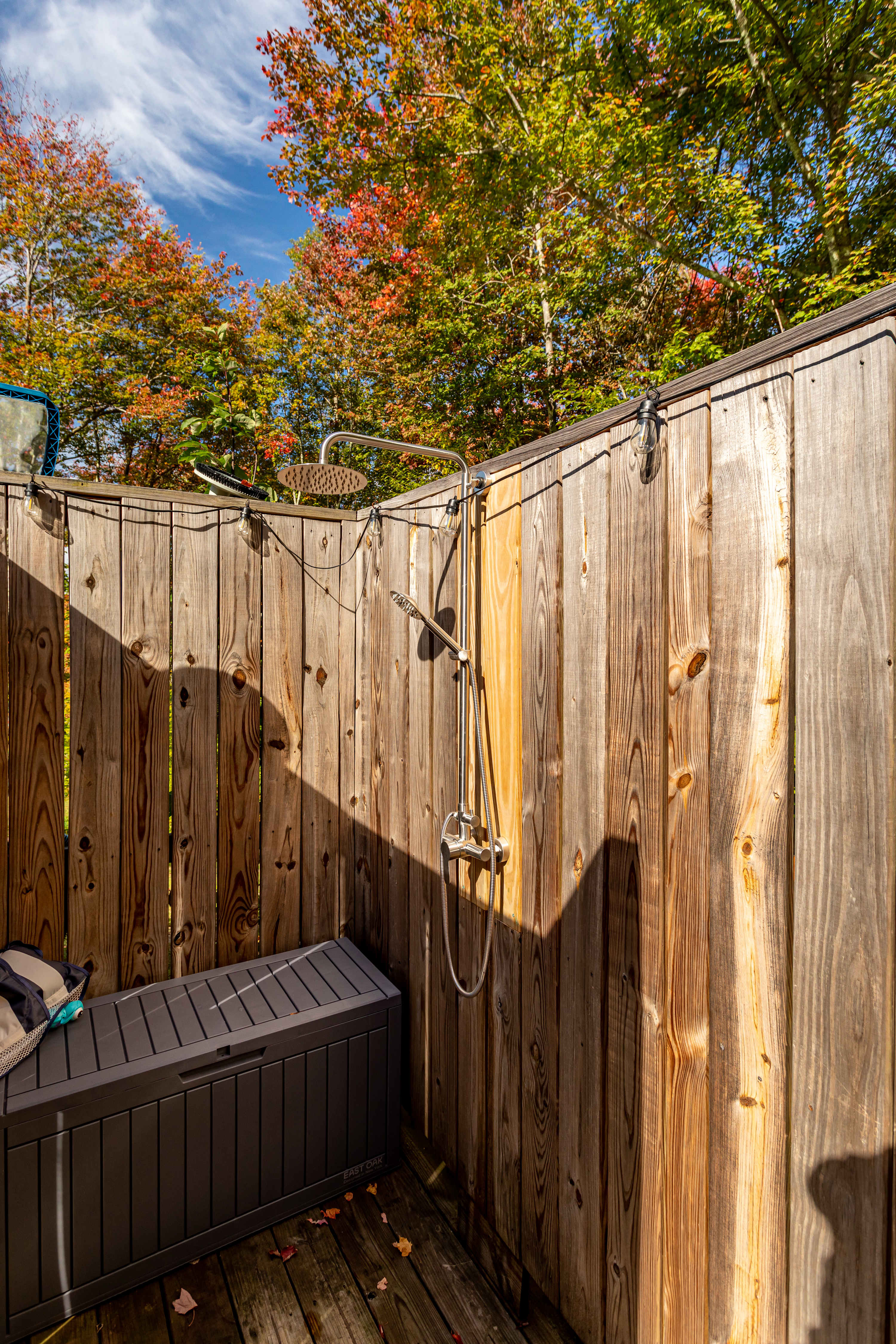 An outdoor shower area enclosed by vertical wooden planks, featuring a modern rain showerhead and a handheld shower attachment. A dark grey storage box sits on the wooden deck floor, with autumn leaves scattered around.