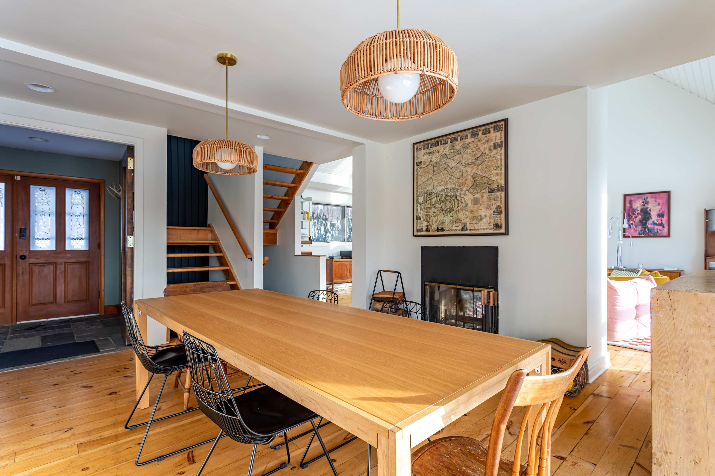A spacious dining area with a long wooden table and a mix of modern and traditional chairs, illuminated by two woven pendant lights. A staircase with wooden treads and a dark accent wall is visible in the background.
