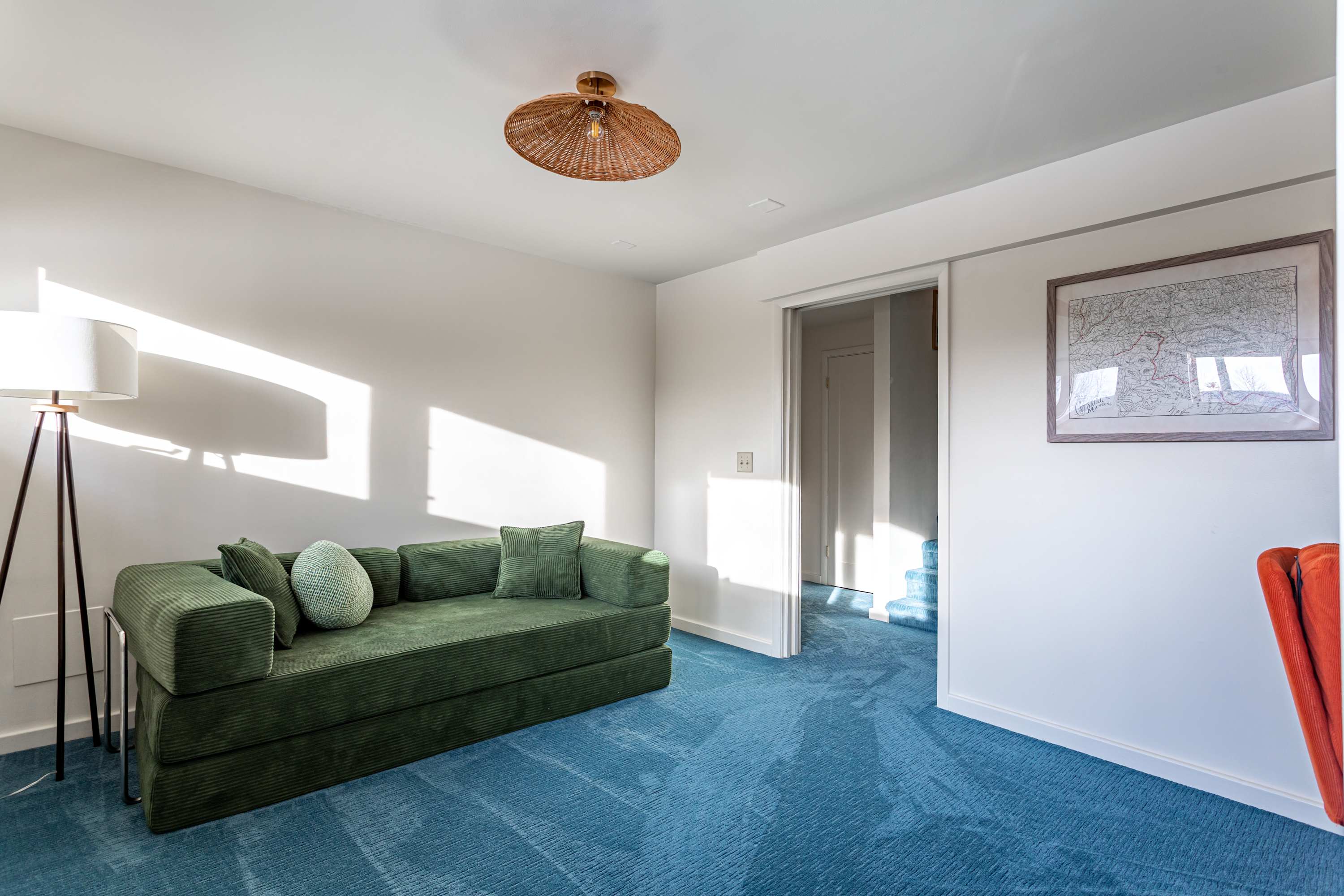 A secondary living area or den with a plush green corduroy sofa and a vibrant blue carpet. Sunlight illuminates the white walls, and a unique woven pendant light hangs from the ceiling, with a doorway leading to another room.
