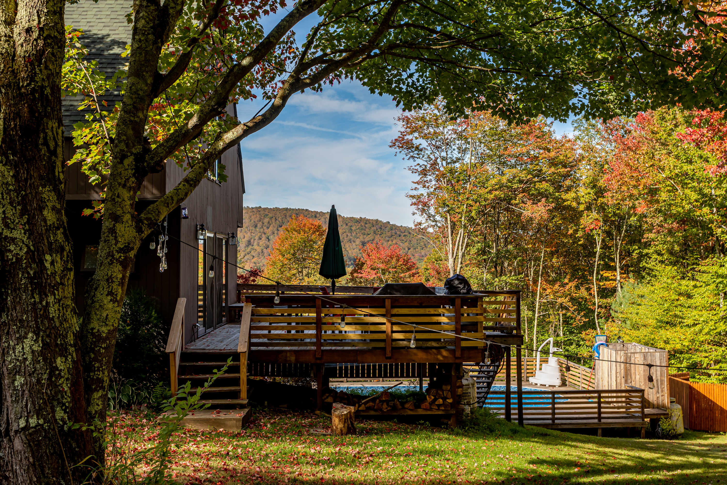 A side view of the brown house with a wooden deck extending from it, featuring a grill and outdoor seating. Below the deck, a swimming pool is visible, all set against a backdrop of vibrant autumn foliage and distant hills.