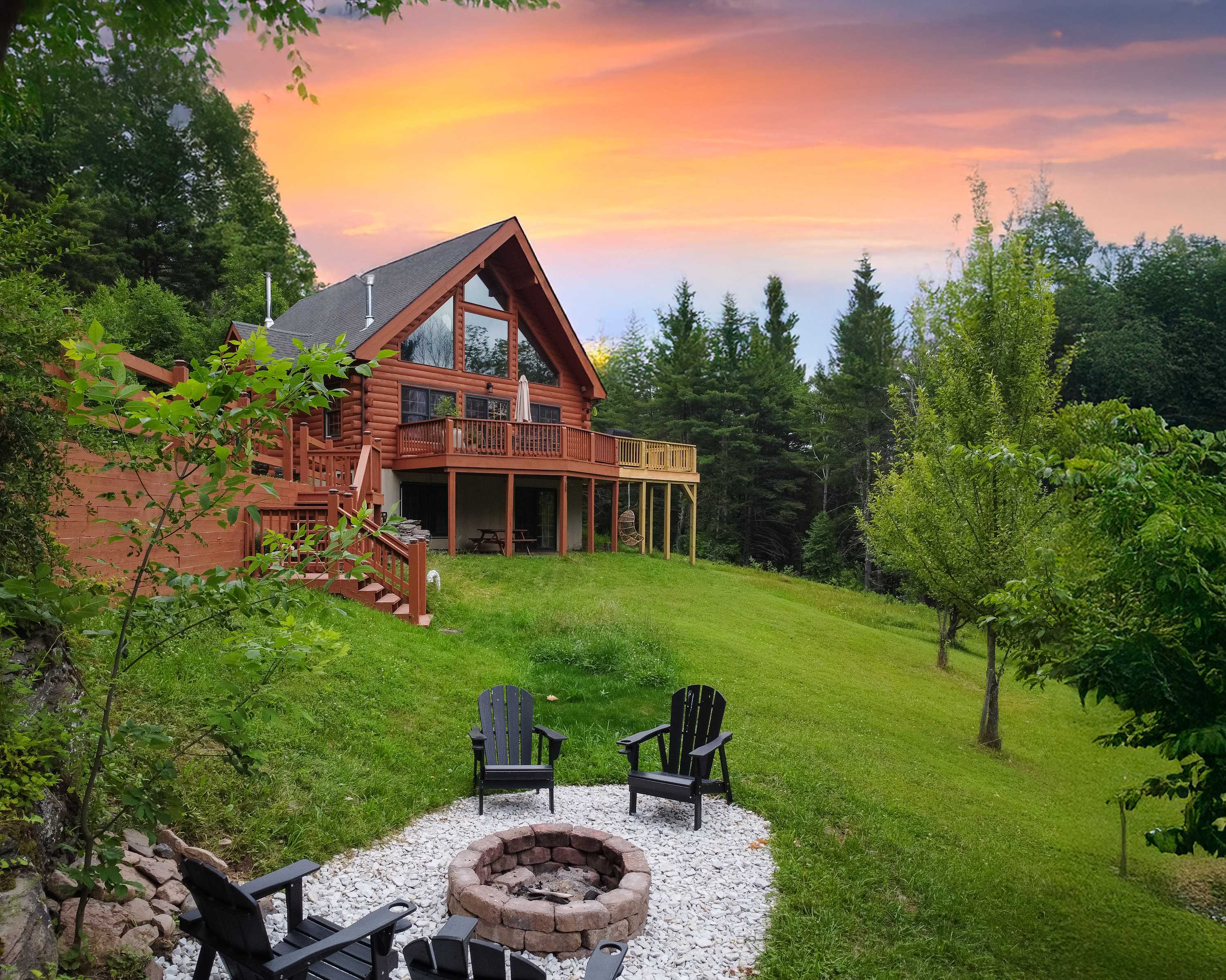 A charming log cabin with a large deck is set on a green hillside, featuring a gravel fire pit area with Adirondack chairs, surrounded by lush trees under a colorful sunset.