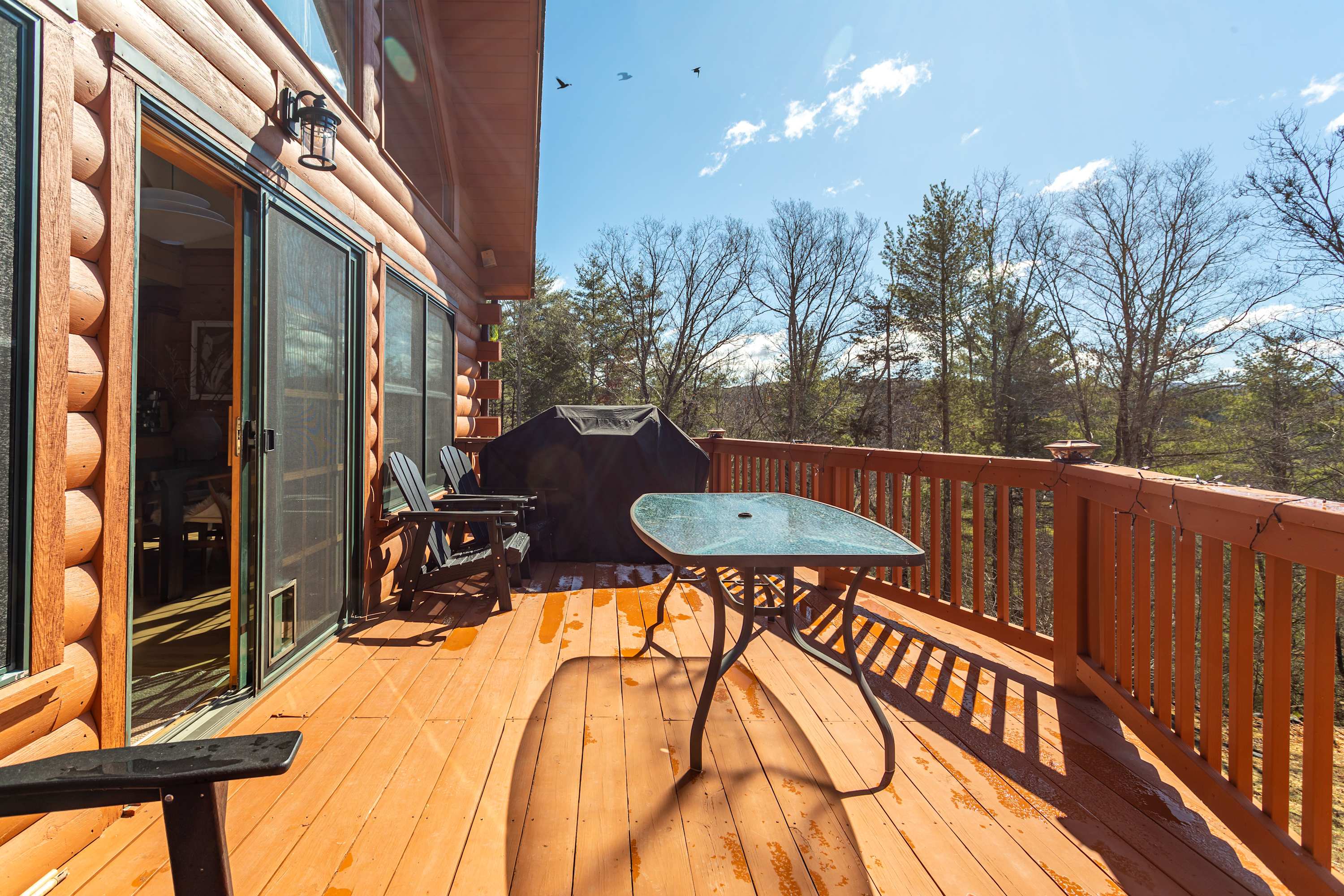 A wooden deck attached to a log cabin, featuring a black BBQ grill and a glass-top table with chairs, overlooking a sunny outdoor landscape with trees.