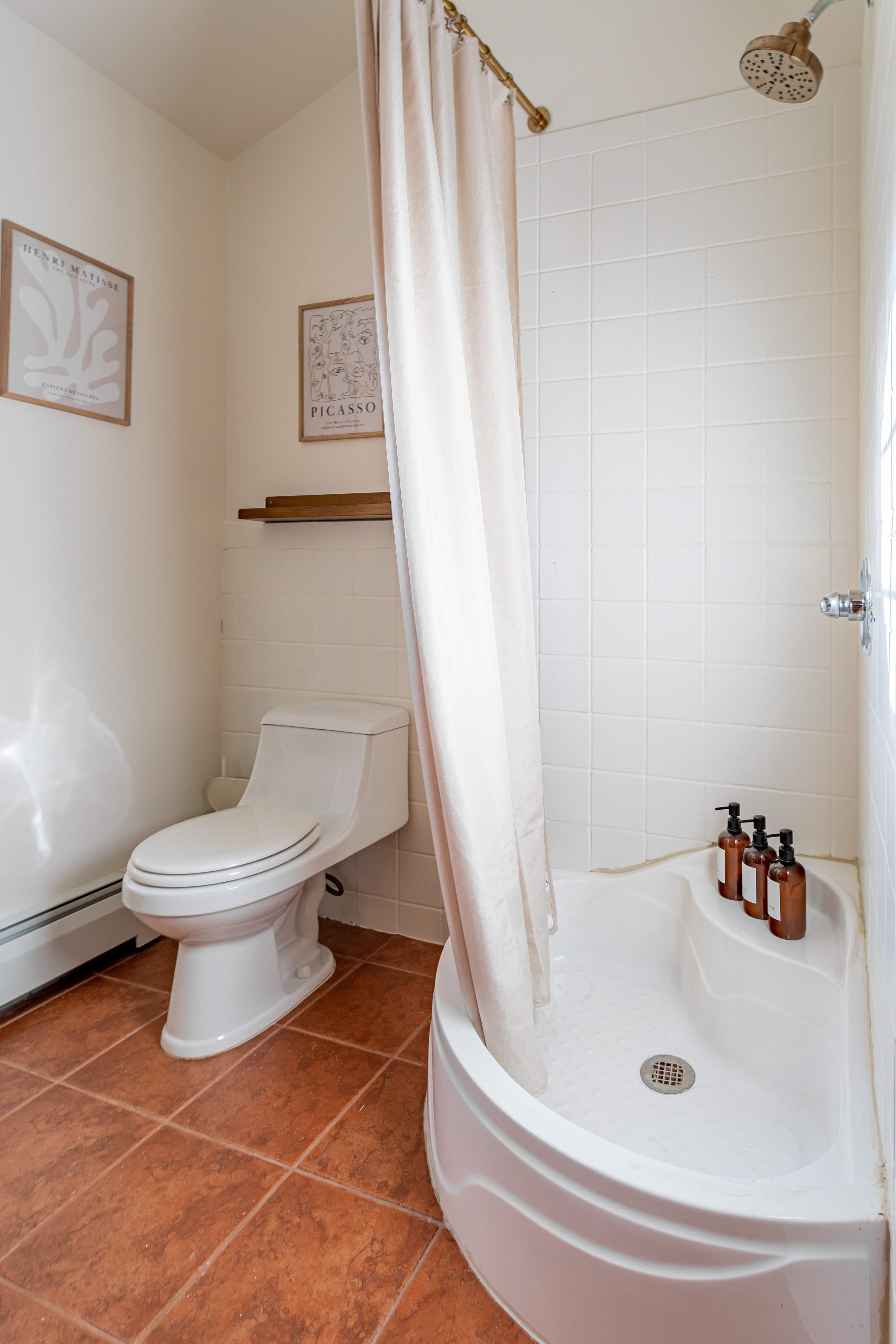 A compact bathroom featuring a corner shower with a white base and tiled walls, a white toilet, and terracotta-colored floor tiles. Two framed art prints adorn the wall above the toilet.