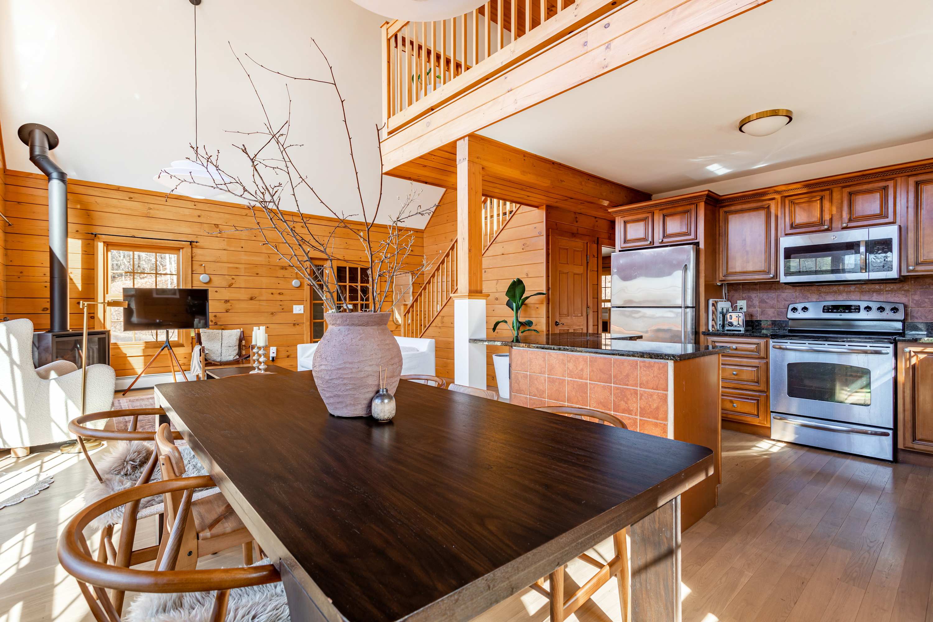 A rustic dining area featuring a dark wood table with modern chairs, set against light wood paneling. The kitchen with wooden cabinets and stainless steel appliances is visible in the background.