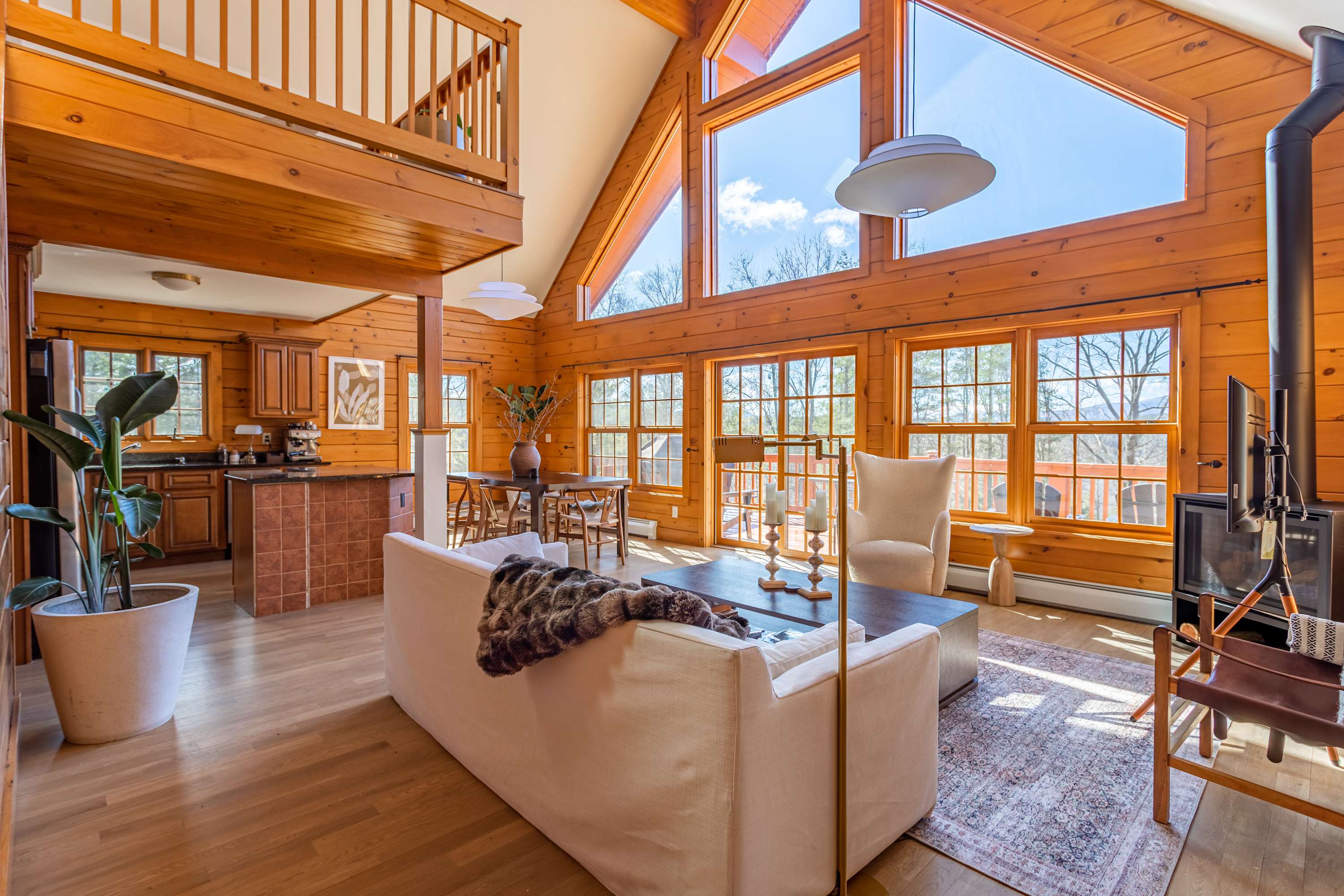 Expansive living space with light wood paneling, a white sofa, and a dark wood coffee table, flowing into a dining area with a large table and views of the deck and mountains through numerous windows.