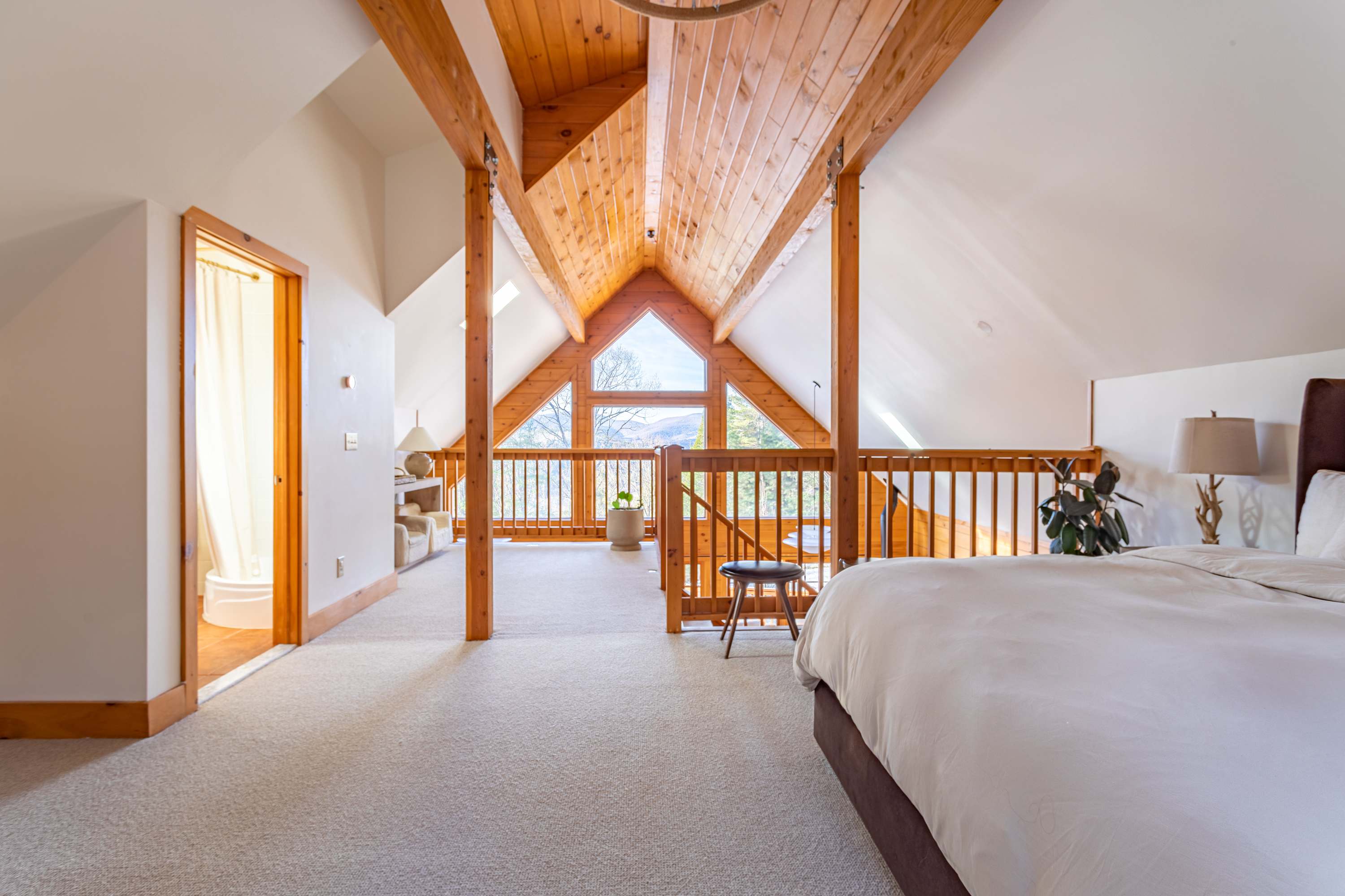A spacious loft bedroom with a vaulted wooden ceiling and exposed beams, featuring a large bed with white bedding, light carpet, and a prominent triangular window offering panoramic mountain views. A wooden railing separates the sleeping area from a lower landing.