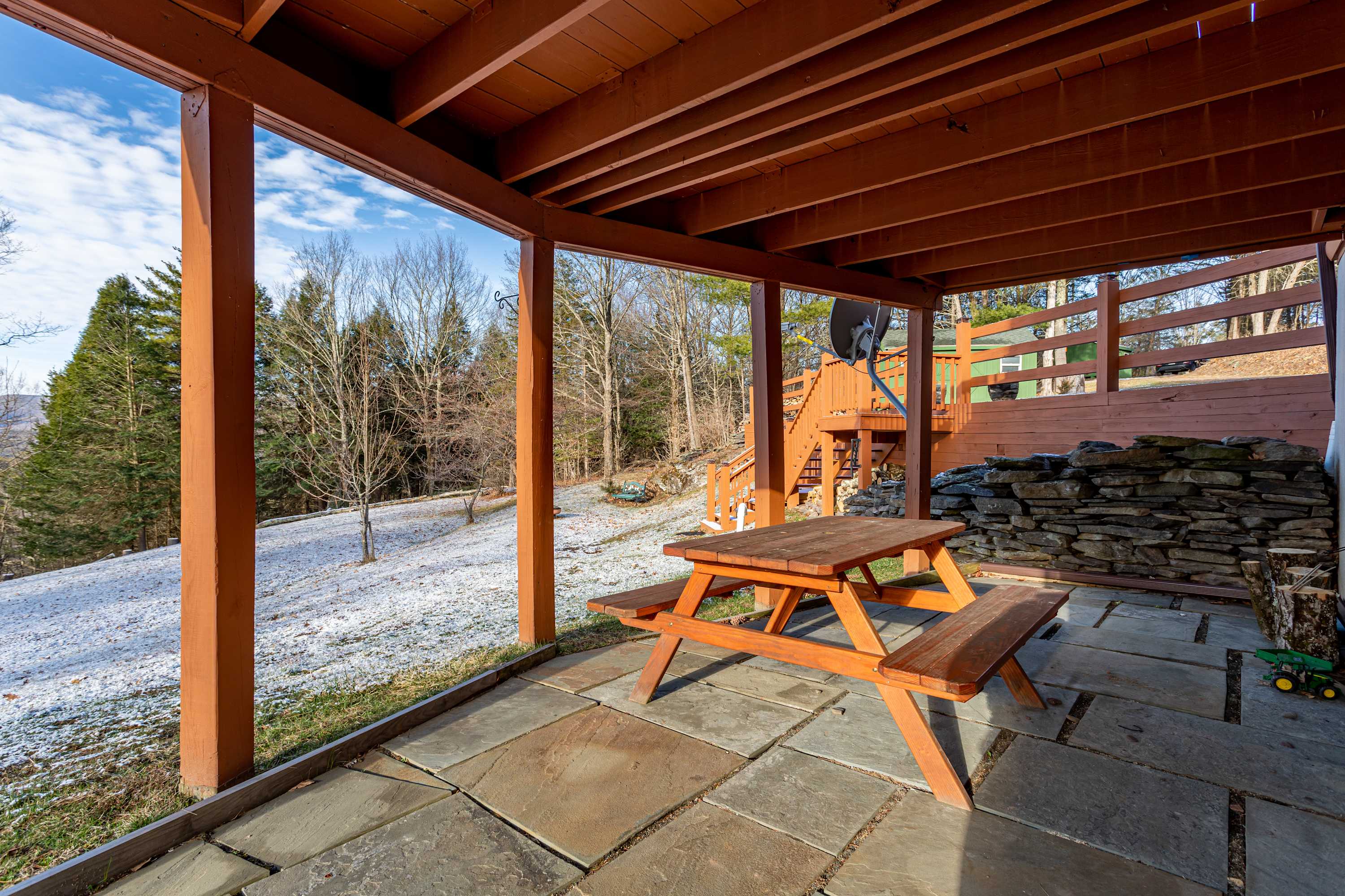 A covered outdoor patio area beneath a wooden deck, featuring a rustic wooden picnic table on large flagstone pavers, with a view of a snowy, wooded hillside.
