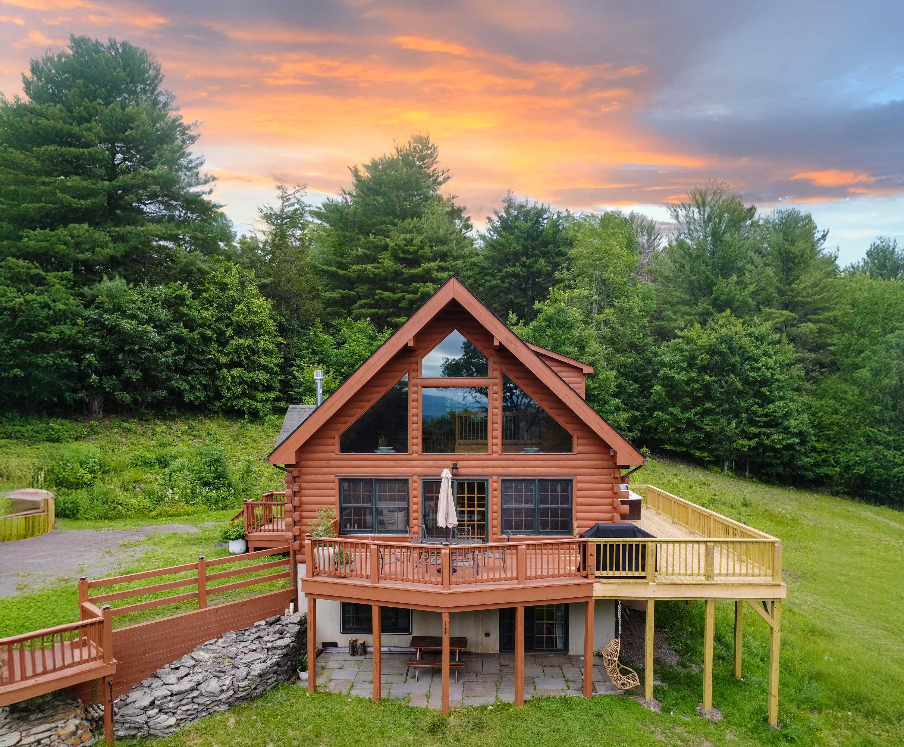 An aerial view of a rustic log cabin with a prominent A-frame and large windows, surrounded by lush green trees and grass under a vibrant sunset sky. A spacious wooden deck with a hot tub and outdoor seating wraps around the house, with a lower patio area featuring a picnic table and hanging chair.