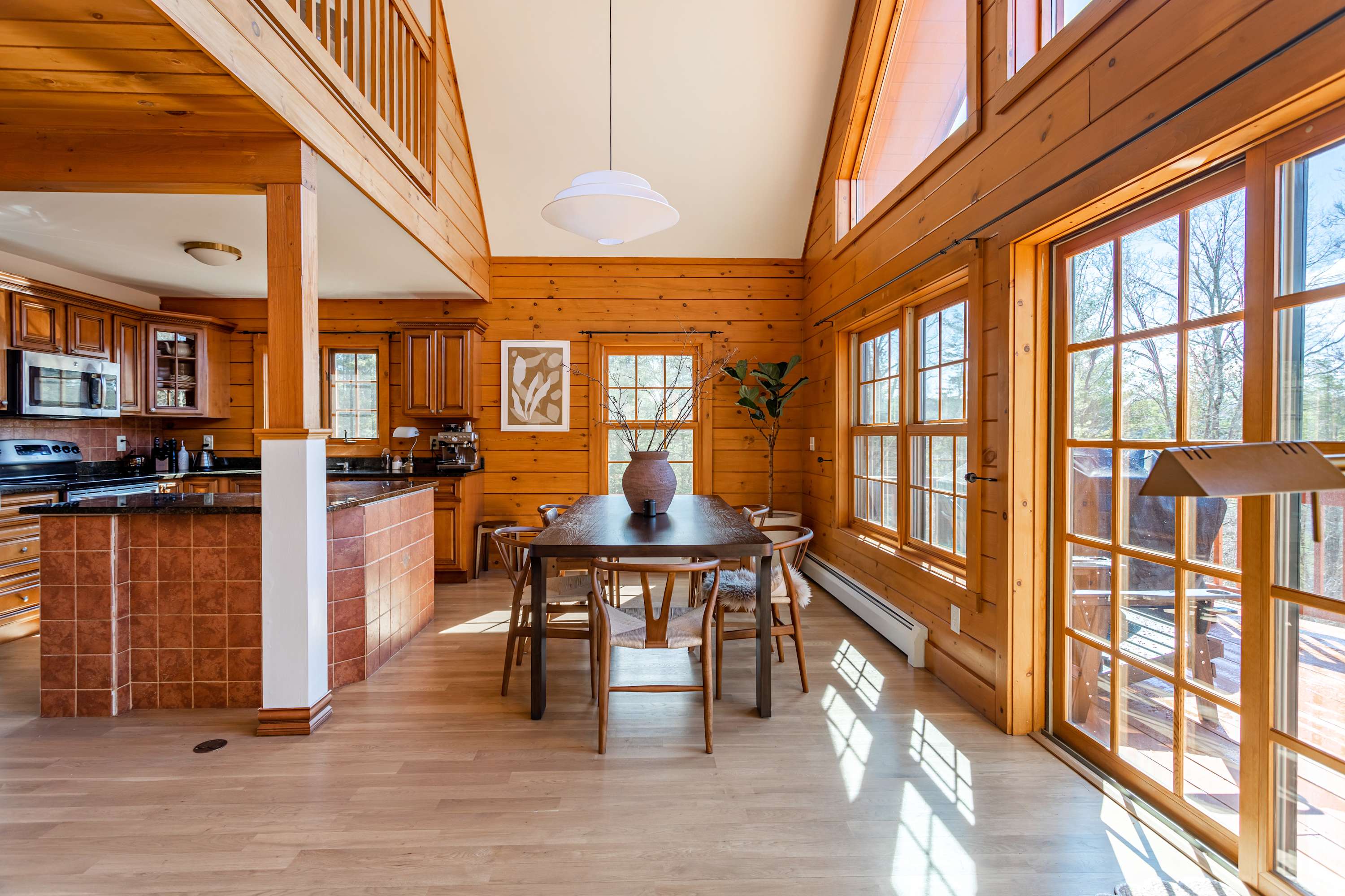 An inviting dining area with a dark wood table and modern chairs, illuminated by natural light from large windows and a glass door leading to an outdoor deck. The kitchen is visible to the left.