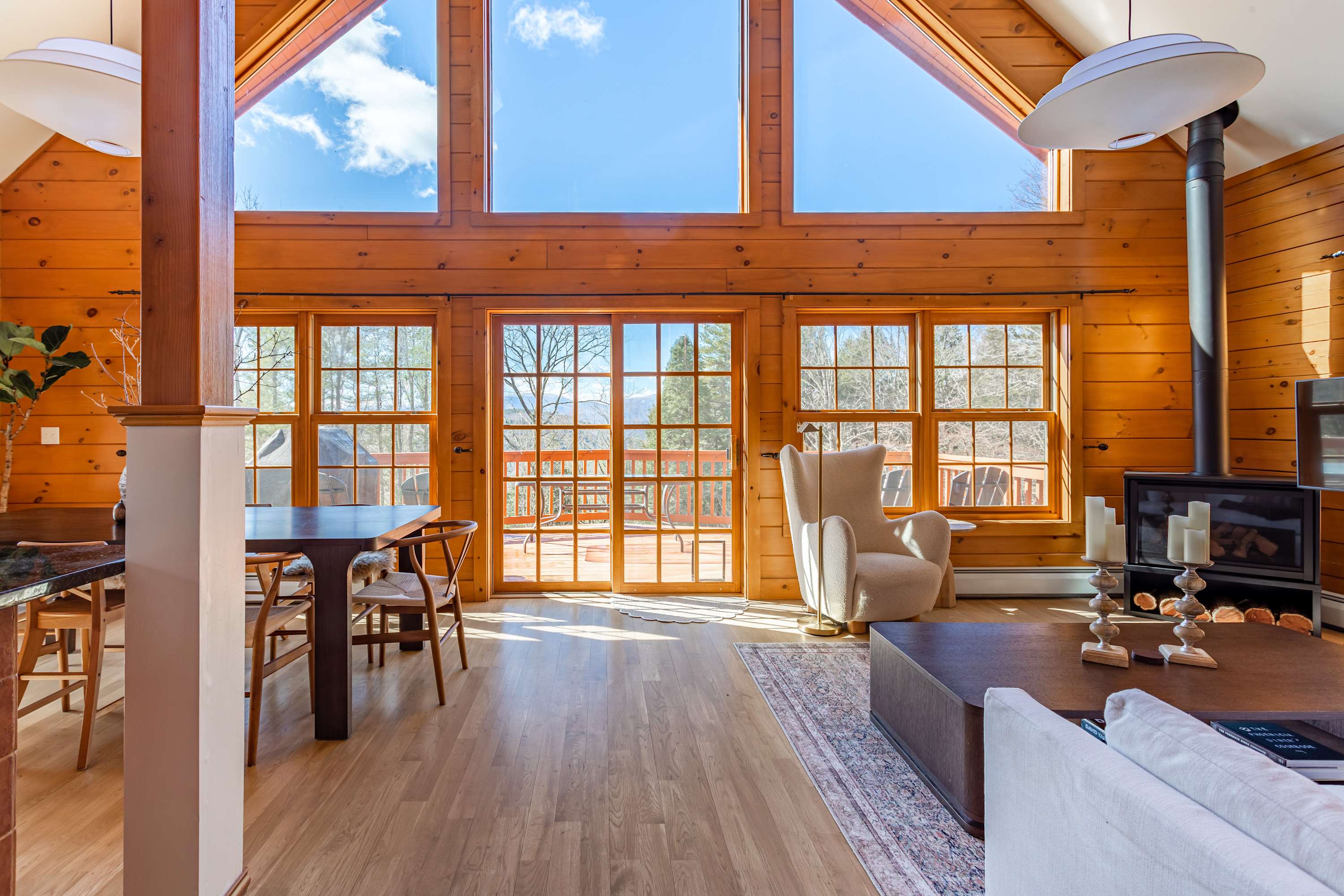 Bright living and dining area featuring light wood paneling, a large dining table, and a cozy armchair, with floor-to-ceiling windows offering extensive views of the outdoor deck and distant mountains.