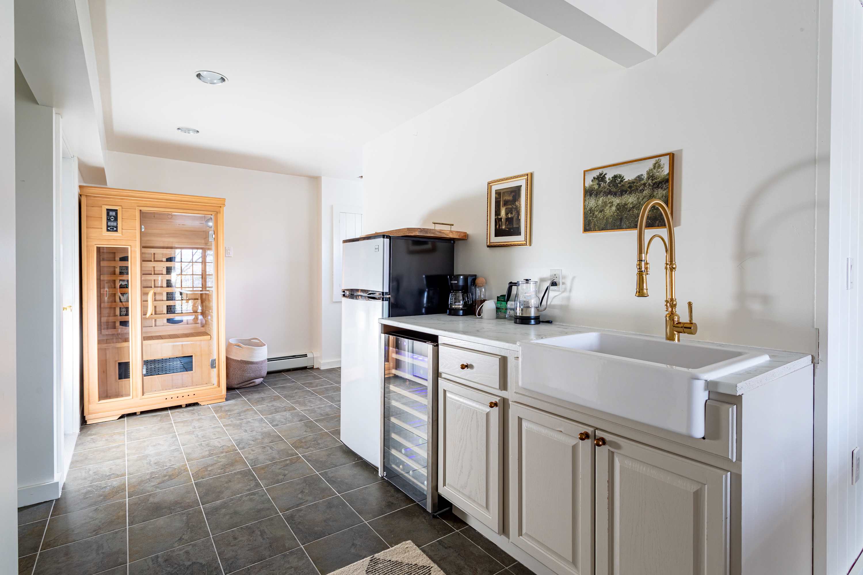 A bright room featuring a light wood sauna with a glass door, alongside a kitchenette area with a white farmhouse sink, gold-toned faucet, light cabinetry, and a mini-fridge with a wine cooler, all on dark tiled flooring.