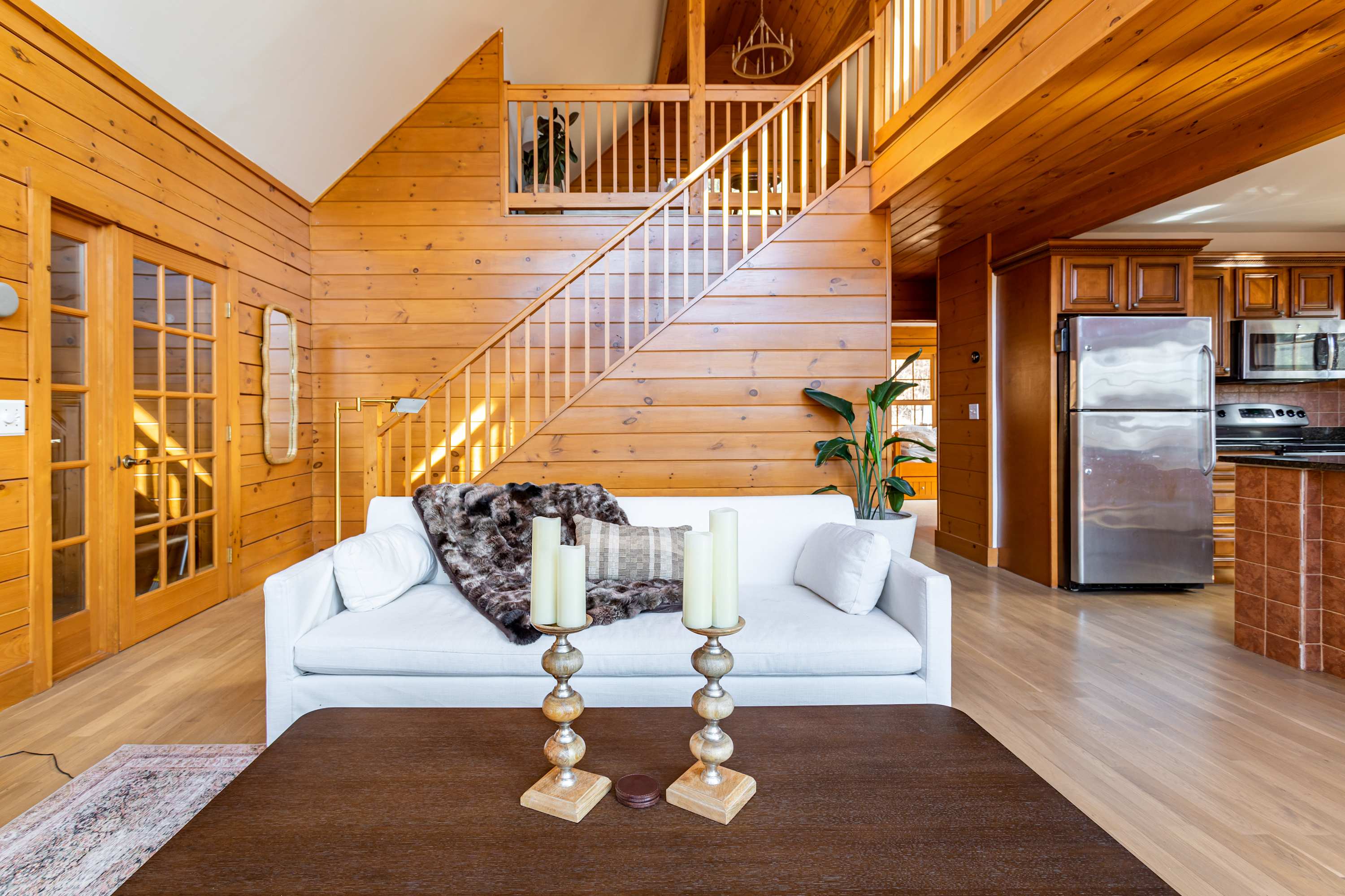 Inviting living room with light wood paneling, a white sofa draped with a faux fur throw, and a dark wood coffee table, with a wooden staircase leading to an upper level.