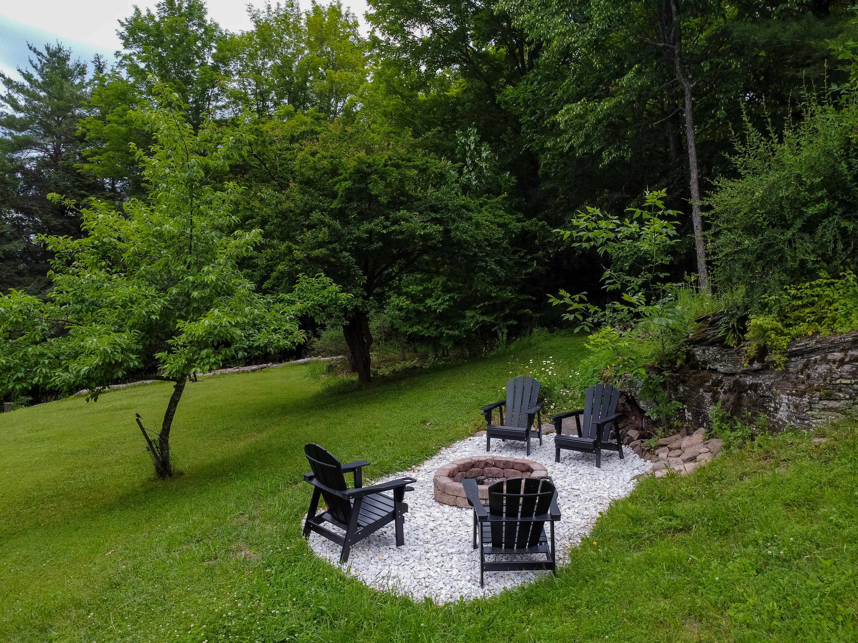 An outdoor fire pit area with a circular stone fire pit surrounded by four black Adirondack chairs, set on a bed of white gravel amidst a grassy lawn and mature trees.