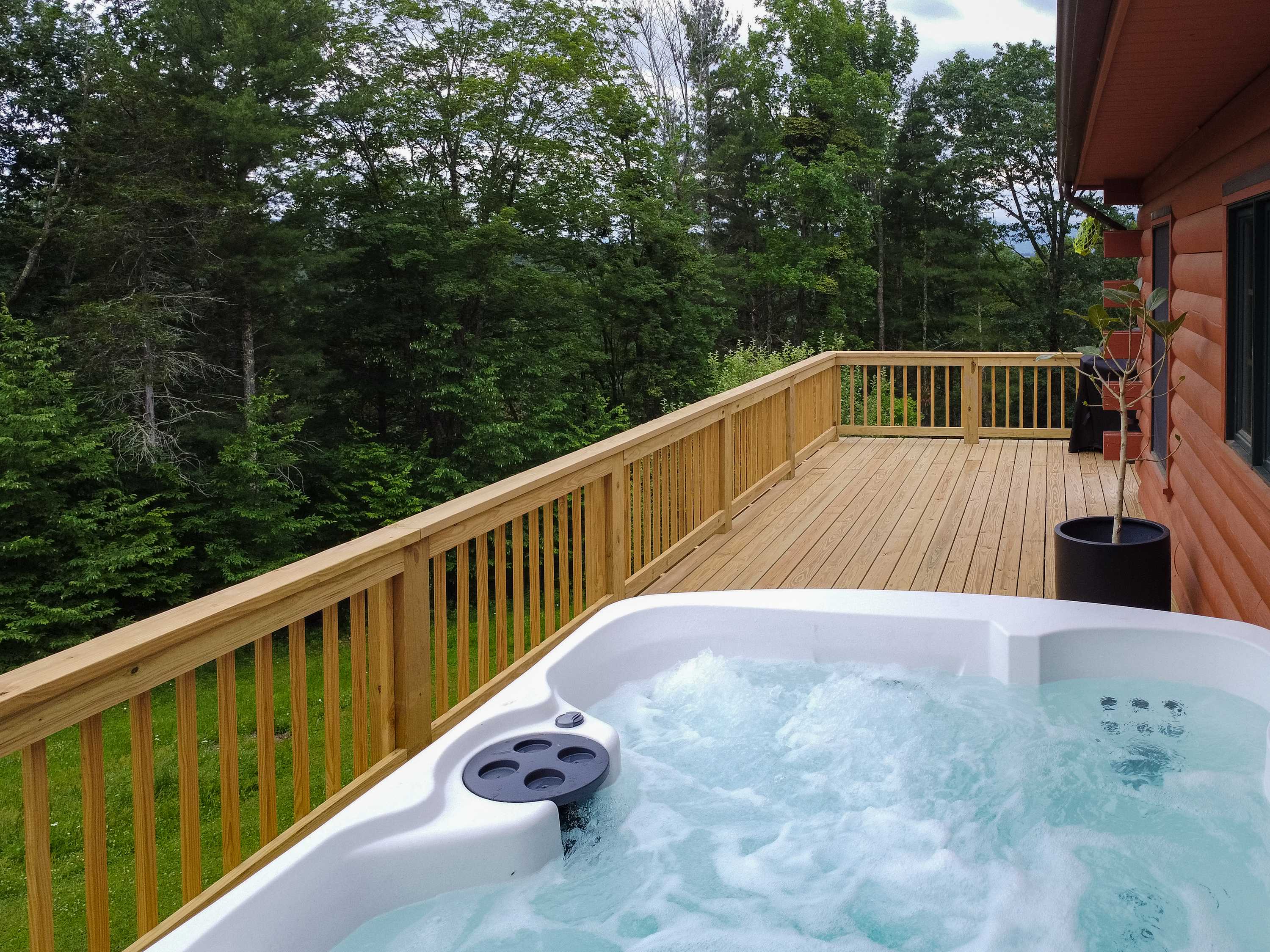 A bubbling white hot tub with black accents is positioned on a spacious wooden deck, offering views of dense green forest in the background.