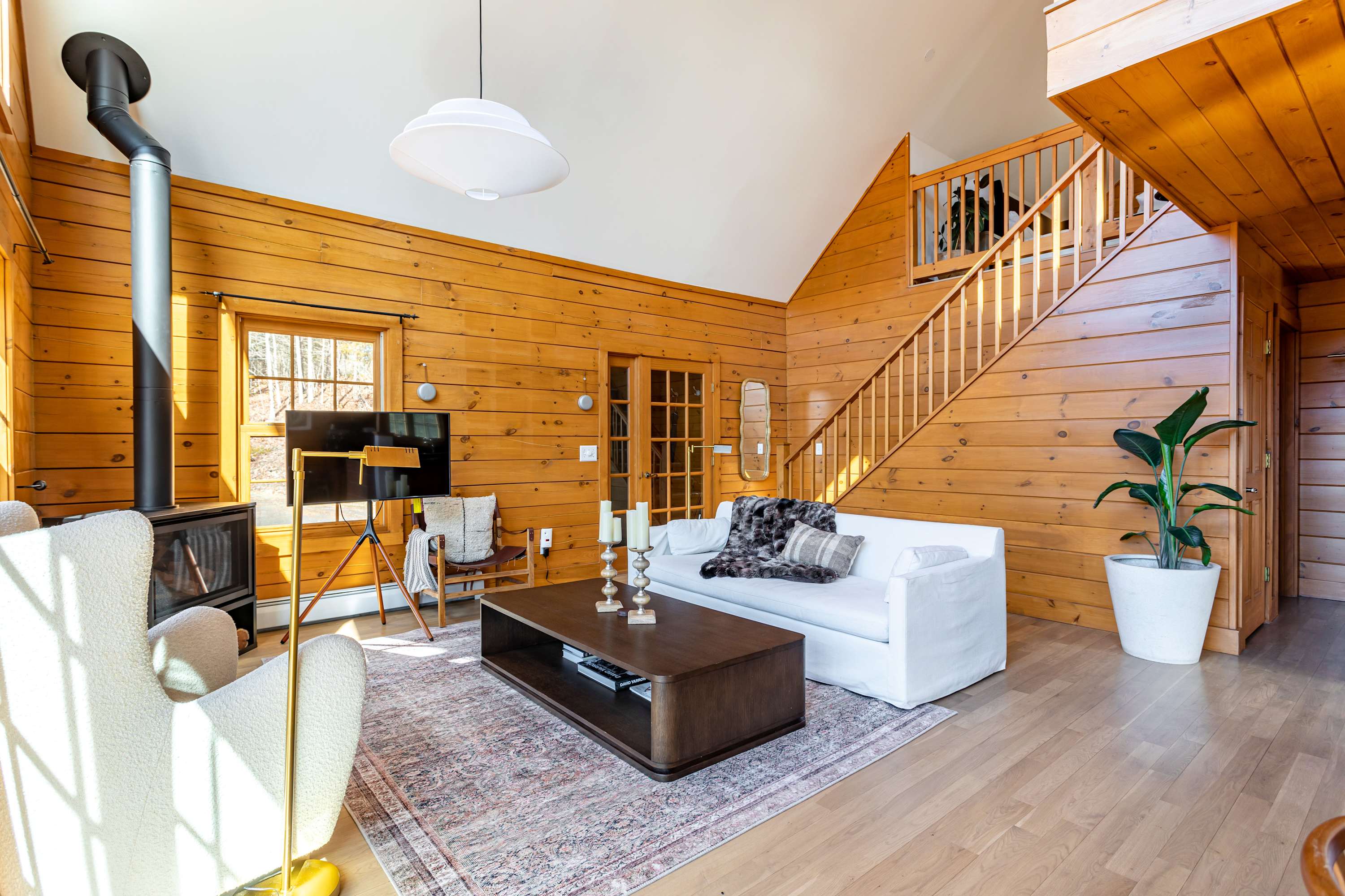 Warm and rustic living room featuring light wood paneling, a white sofa, a modern wood-burning stove, and a wooden staircase leading to the upper floor, all illuminated by natural light.