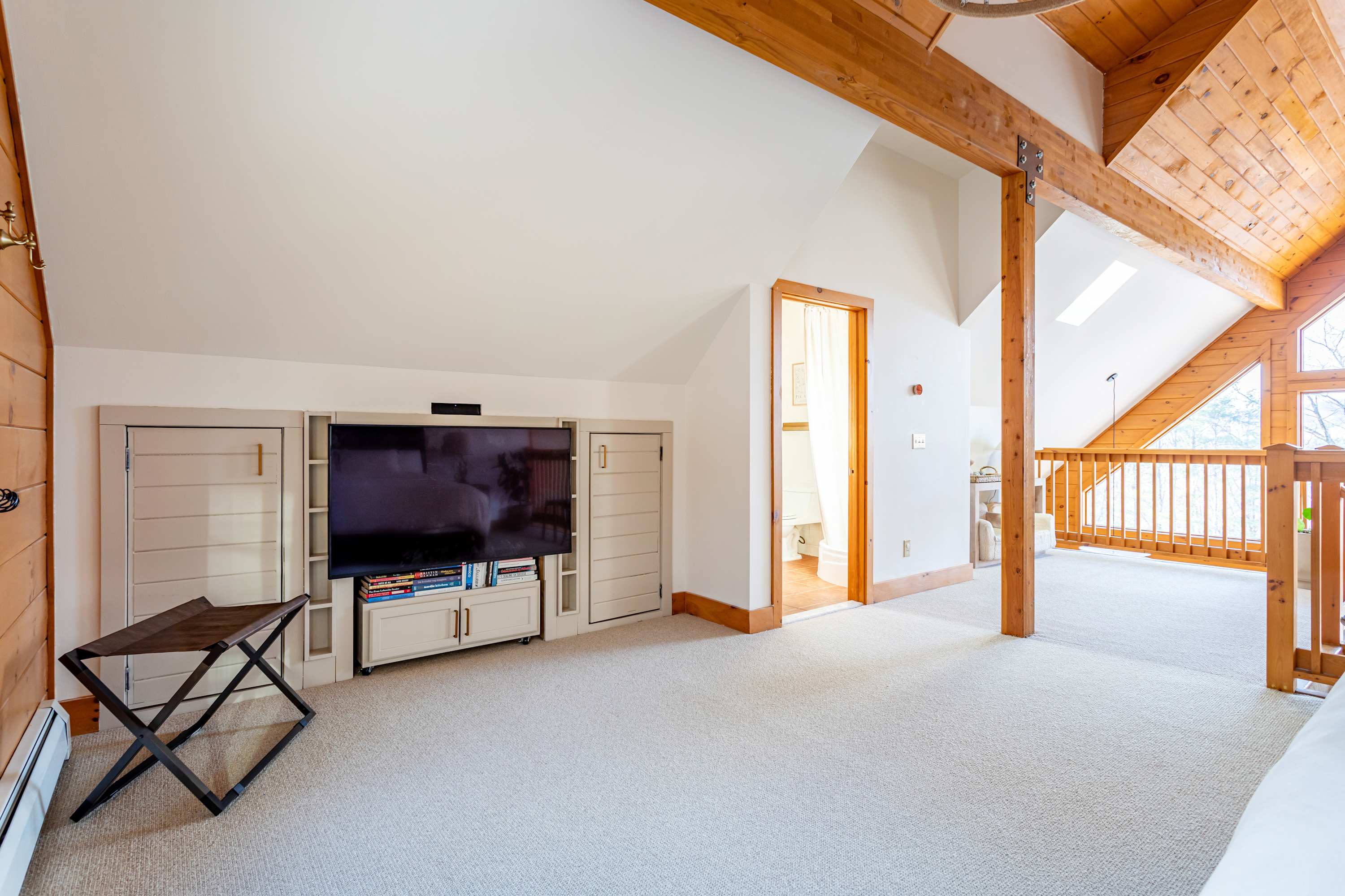 A bright loft area with light carpet, built-in cabinetry housing a large TV, and an open railing overlooking the main living space. A doorway leads to another room.