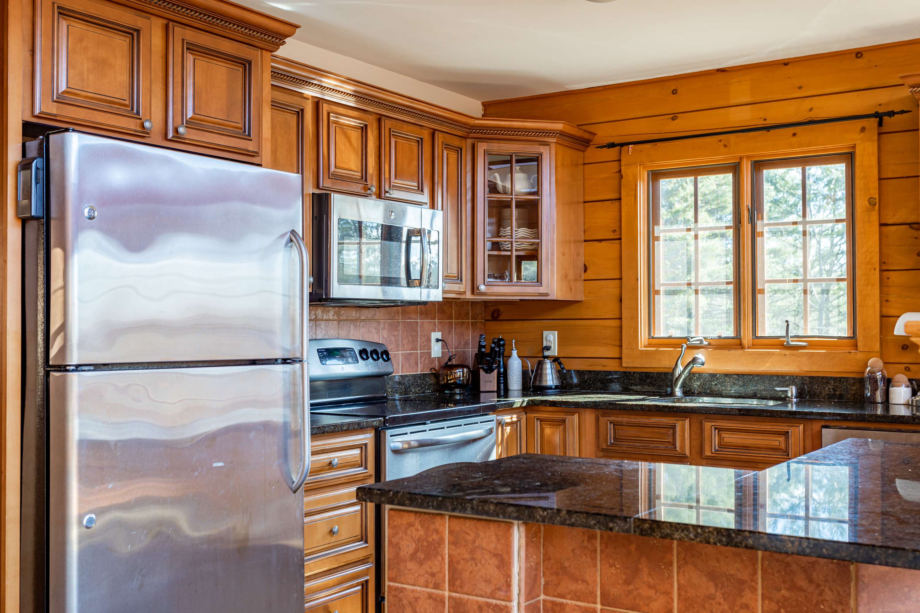 A well-equipped kitchen with warm wooden cabinets, dark granite countertops, and stainless steel appliances including a refrigerator, microwave, and stove. A window overlooks the outdoors.