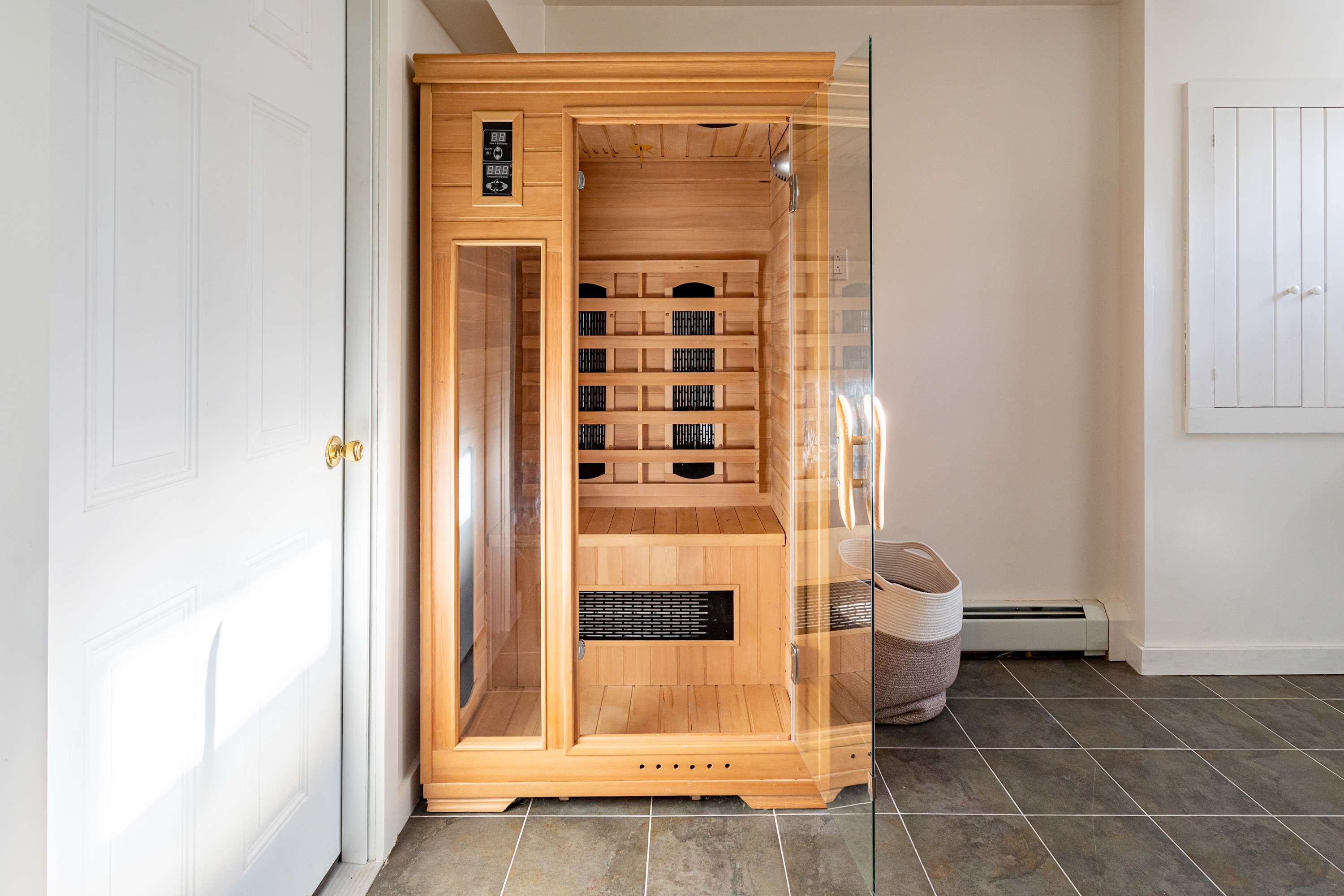 A modern wooden infrared sauna with a glass door, located on a tiled floor next to a white door. A woven basket is placed beside the sauna.