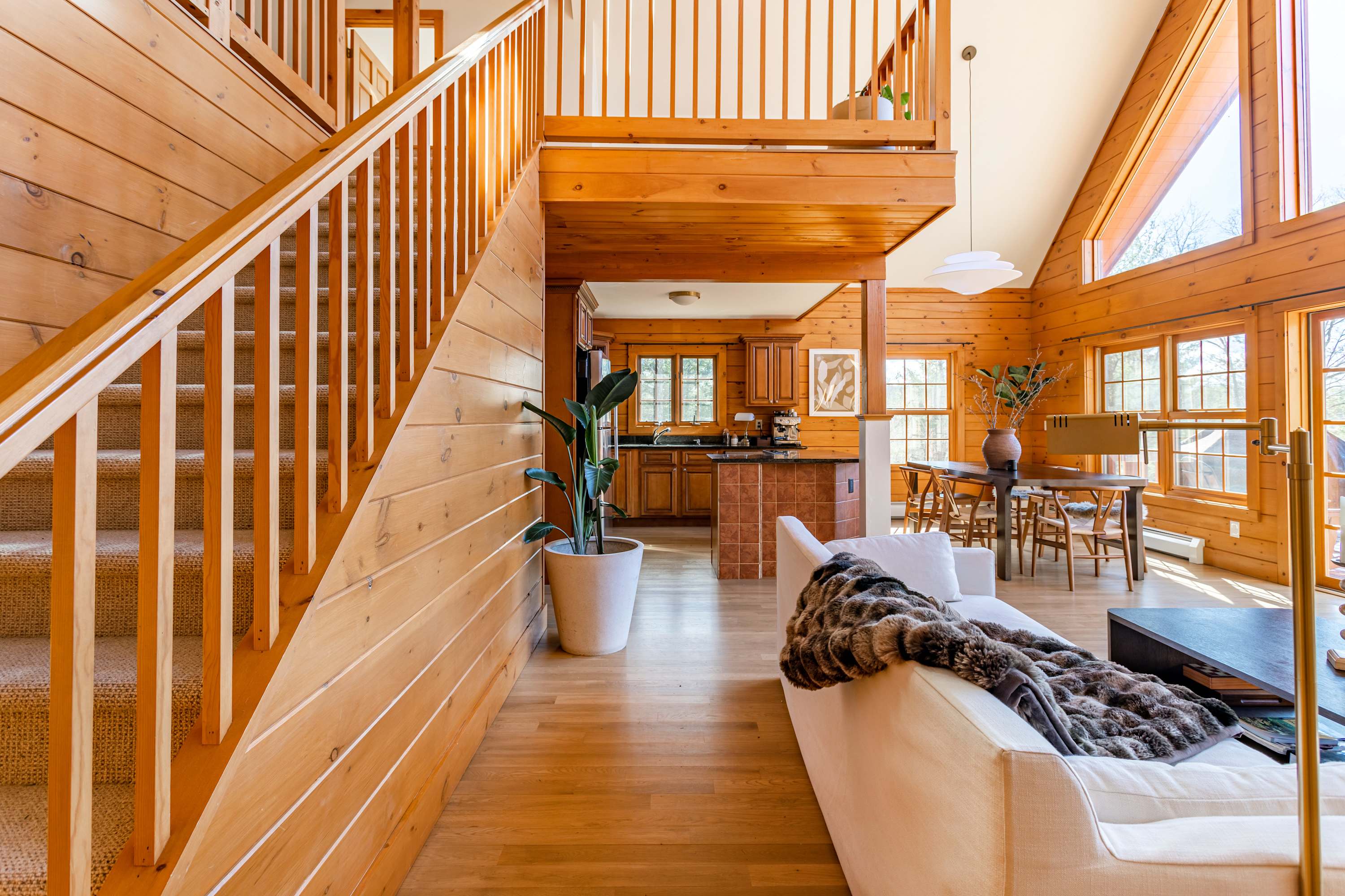 An expansive open-concept living space with light wood paneling, a white sofa, and a dining area. A prominent wooden staircase leads to the upper level, showcasing the chalet's rustic charm.