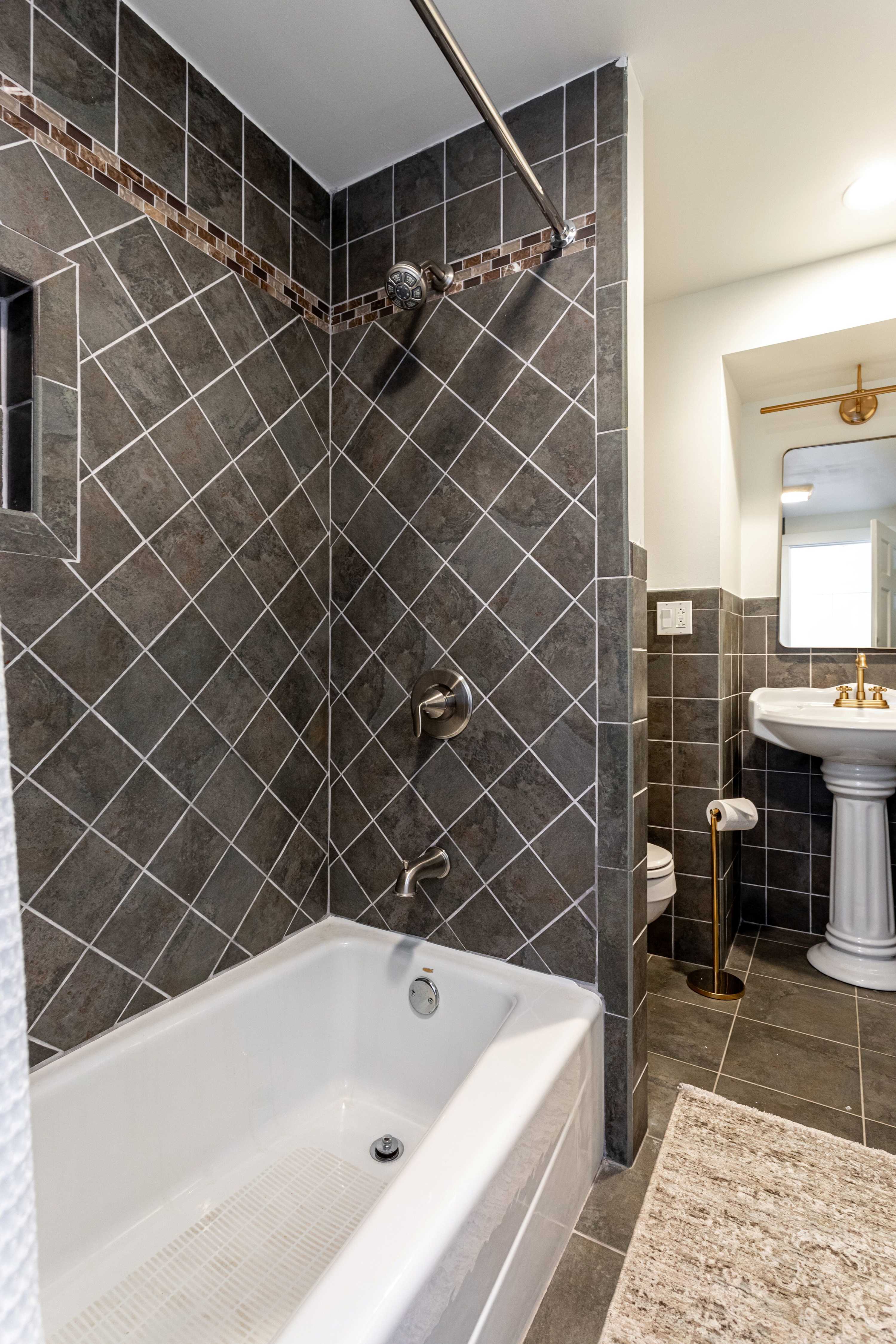A shower-tub combination in a bathroom with dark gray diagonal tiles on the walls and a decorative mosaic border. A brushed nickel shower head and faucet are installed, with a white tub below.