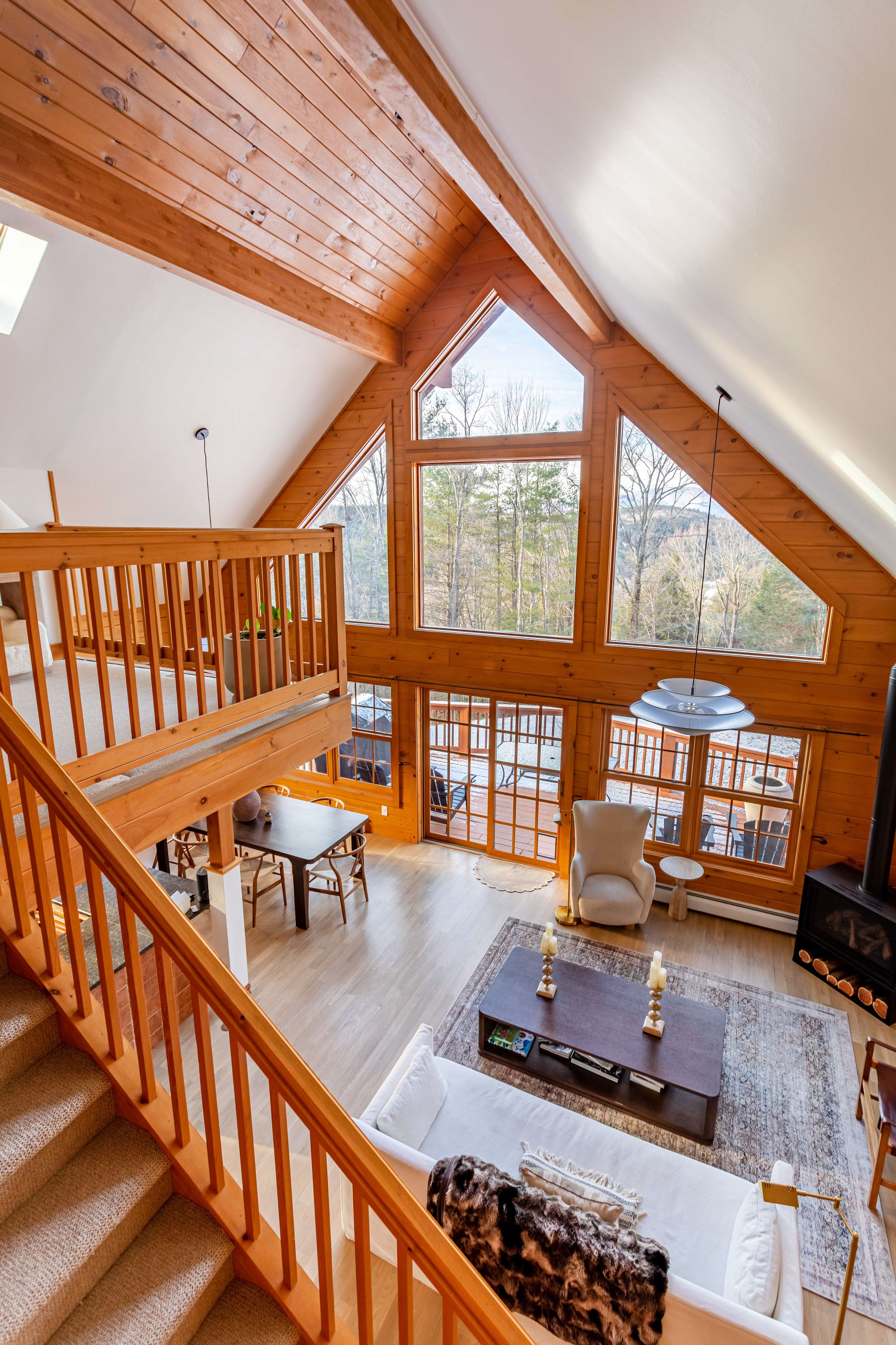 A high-angle view of the open-concept main floor, showcasing the living room with a sofa and fireplace, and the dining area. A wooden balcony overlooks the space, with large windows offering mountain views.