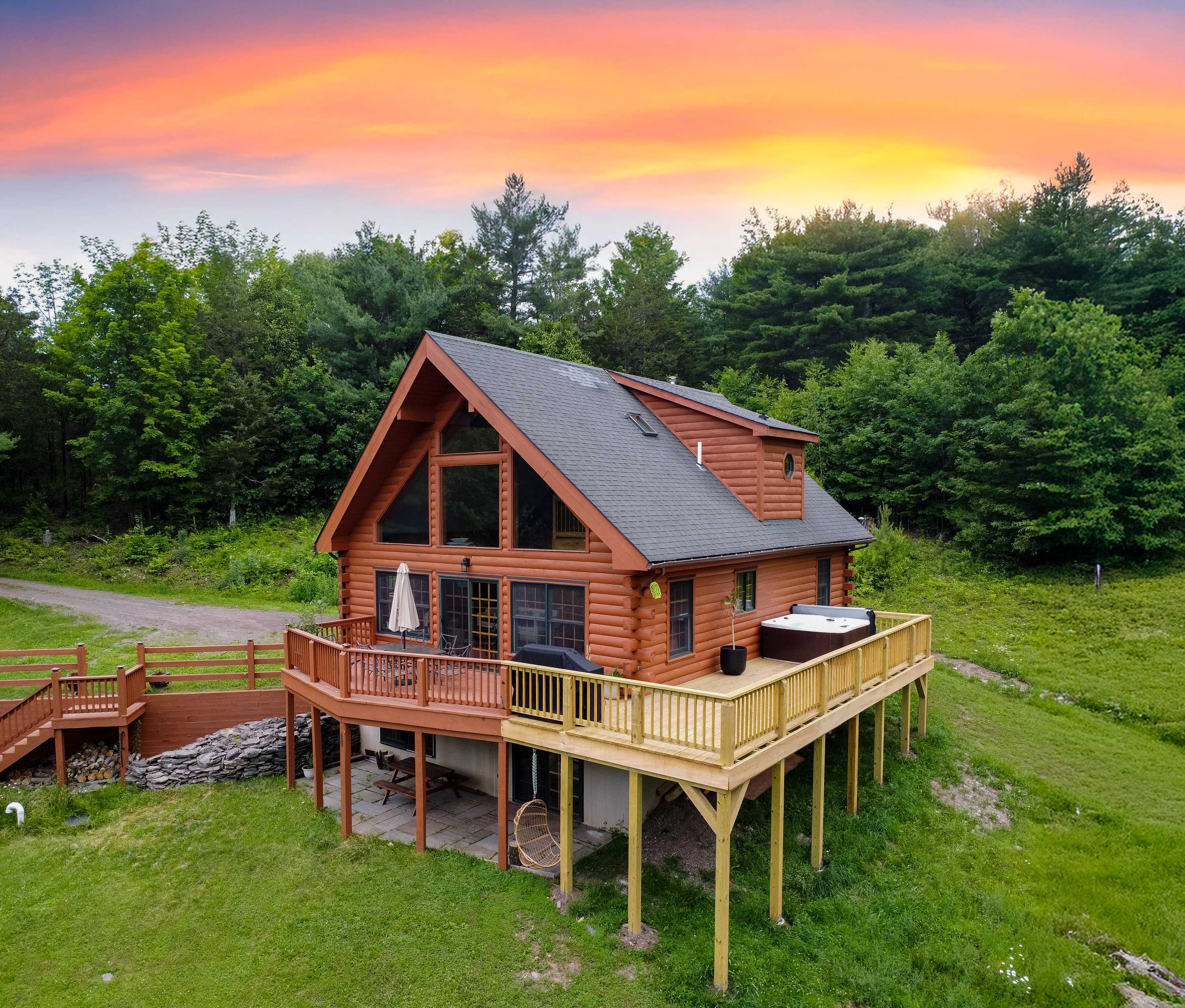Aerial view of a rustic log cabin with a dark roof and a large wooden deck, nestled on a grassy hillside surrounded by dense green trees under a vibrant sunset sky.