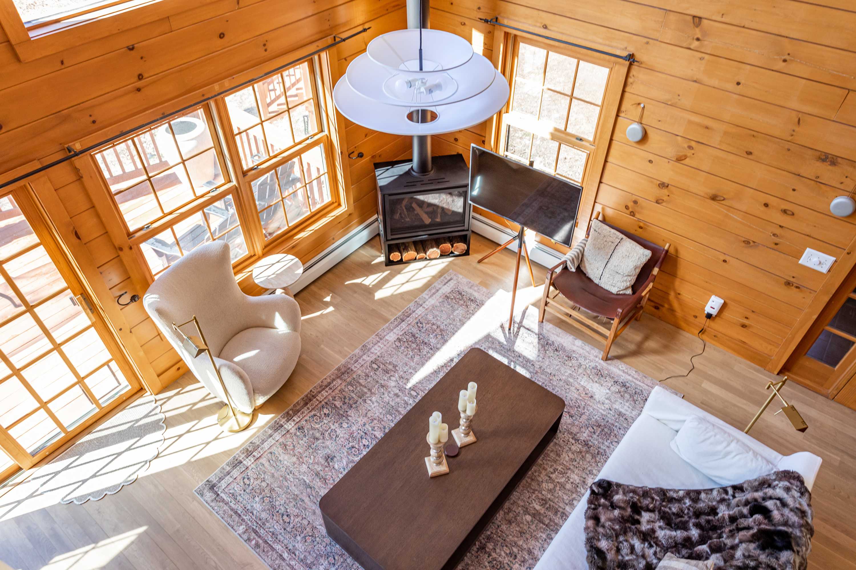 An overhead view of the living room featuring a white sofa, dark coffee table, and a modern fireplace. Large windows and a high ceiling create a bright and airy atmosphere.